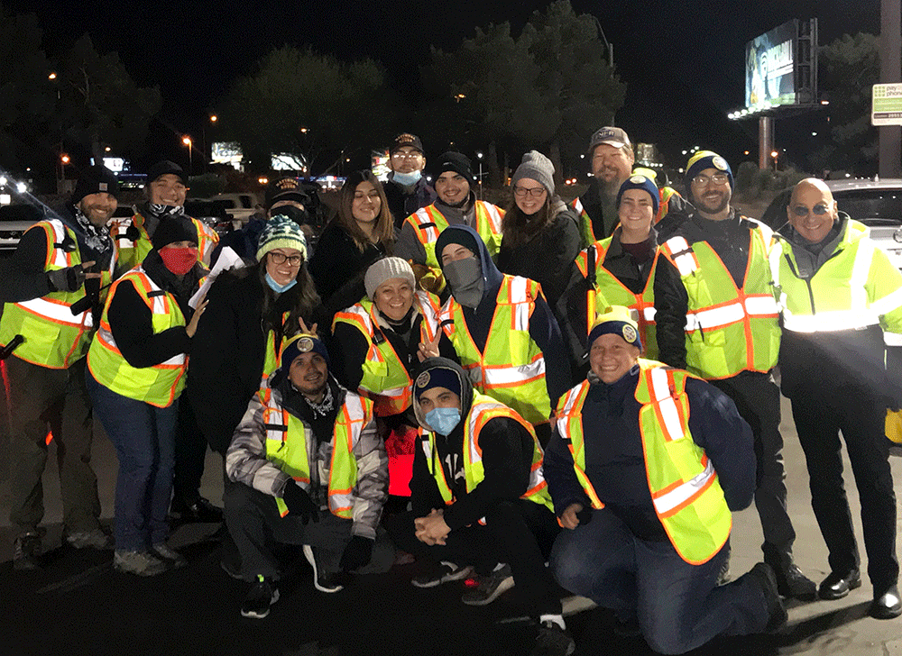 Group of people wearing reflective safety vests gathered outdoors at night, smiling for the camera.