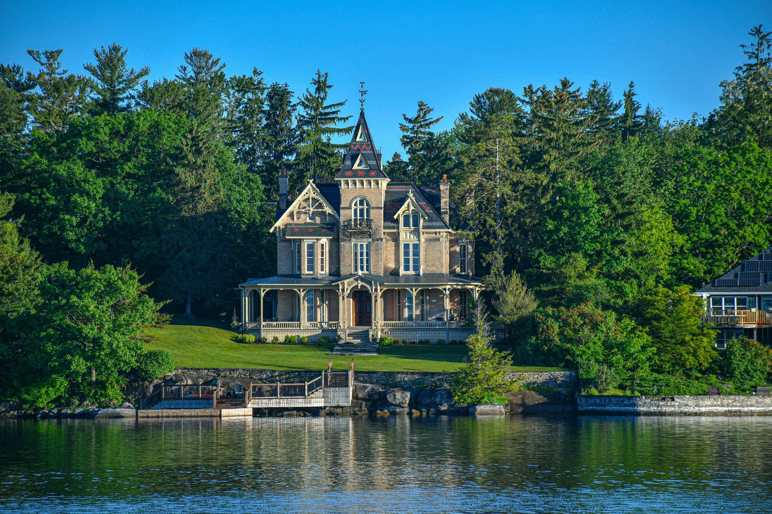 A large Victorian-style house with a steeply pitched roof and decorative trim, situated at the edge of a lake, surrounded by tall green trees, under a clear blue sky.