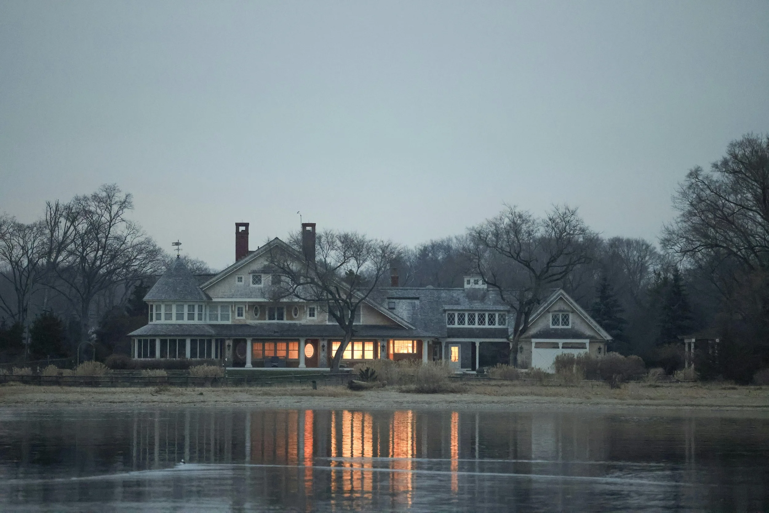 Large house with numerous windows and chimneys, illuminated from within, located by a lake reflecting its lights, surrounded by leafless trees under a cloudy sky.
