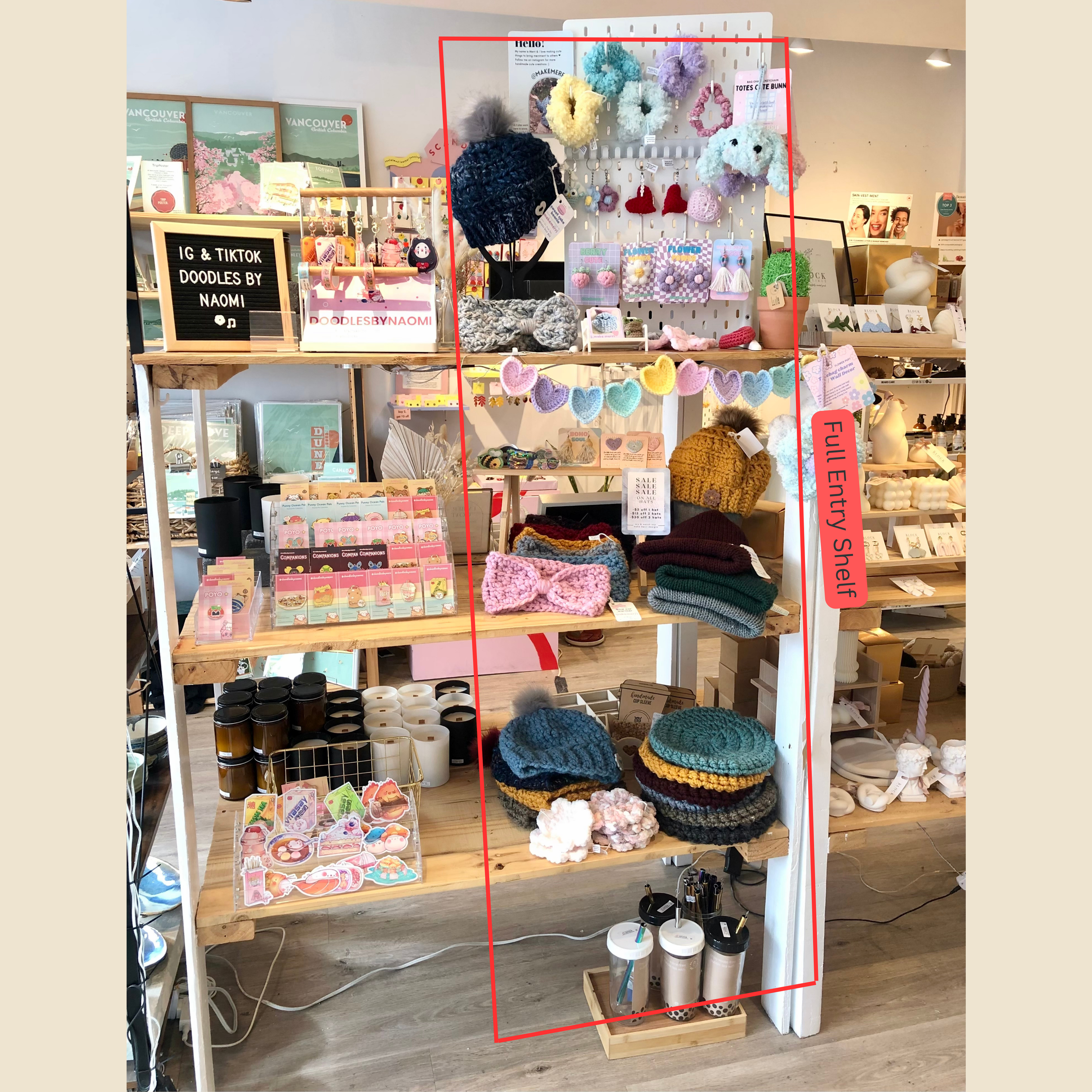 A retail display shelf filled with colorful handmade knit hats, scarves, and plush accessories, with a sign indicating it's a full entry shelf at a boutique shop.