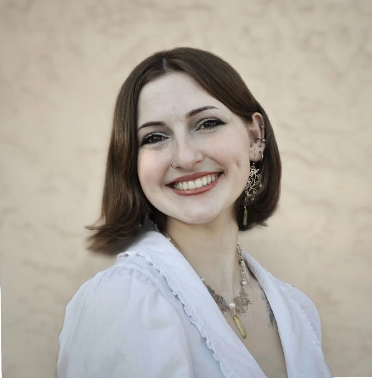 A smiling woman with shoulder-length brown hair, wearing a white blouse and jewelry, standing against a beige background.