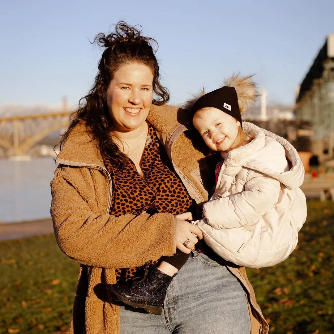 A woman smiling and holding a young girl in her arms outdoors near a river, with a bridge in the background on a sunny day.