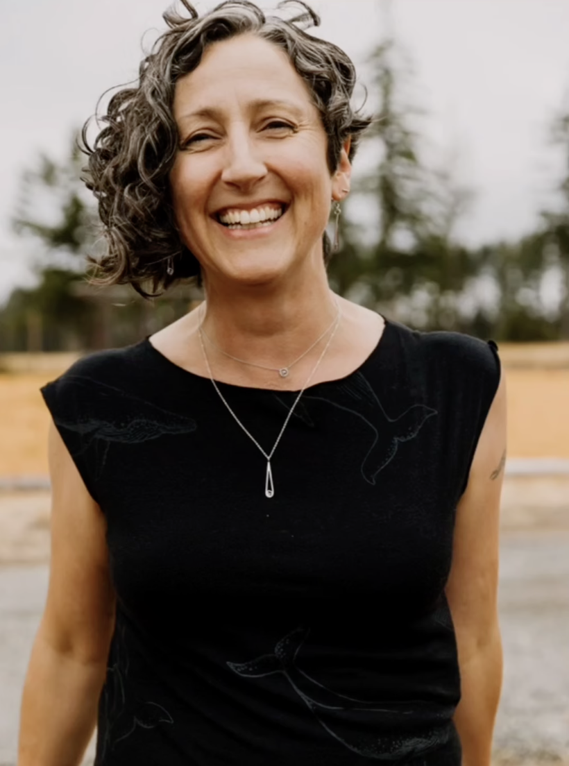 A smiling woman with short, curly gray hair, wearing a black sleeveless top with a subtle feather pattern, and a delicate necklace, standing outdoors with a blurred background of trees and sky.
