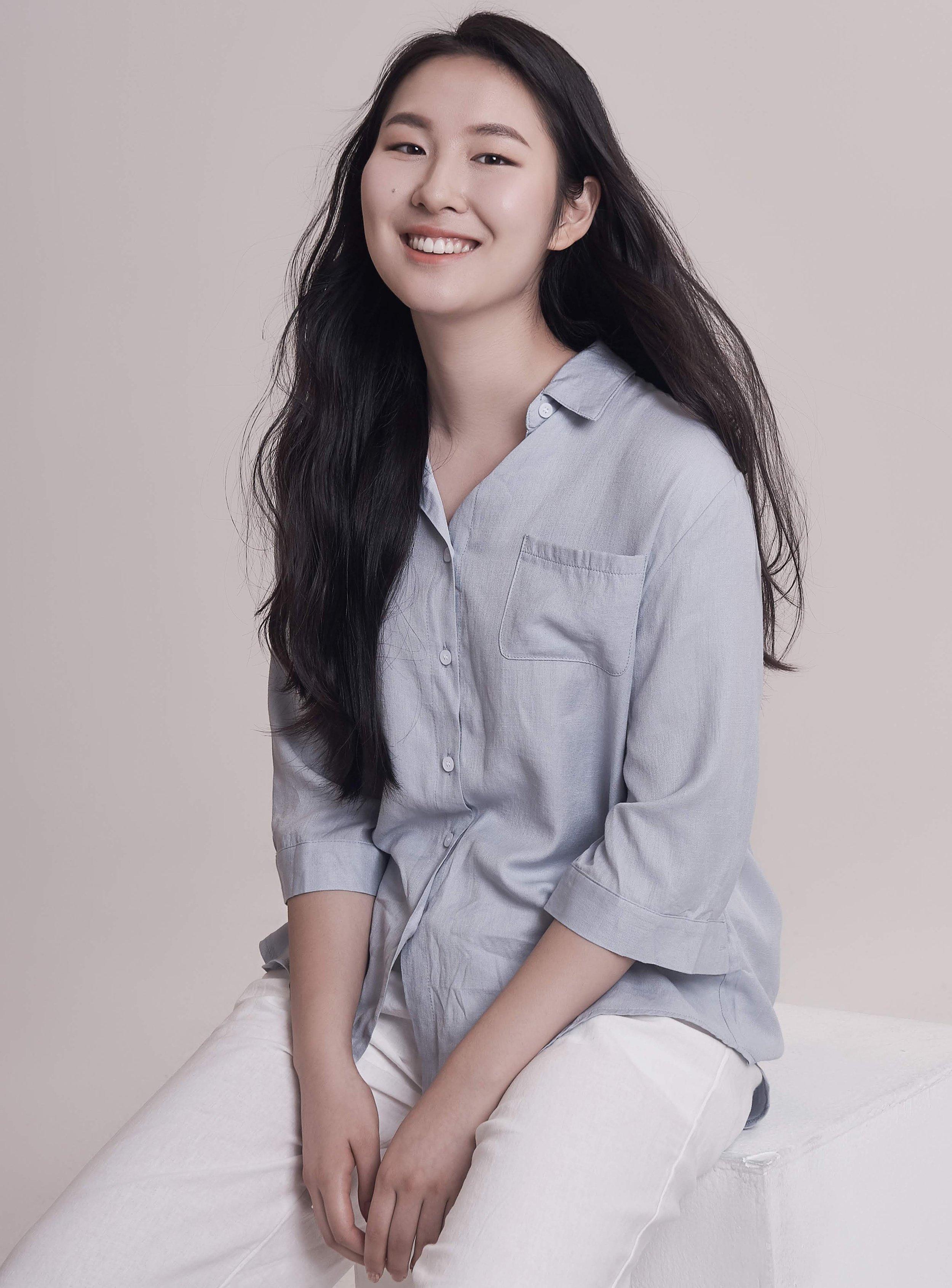 A young woman with long black hair, smiling, sitting on a white surface, wearing a light gray button-up shirt and white pants.