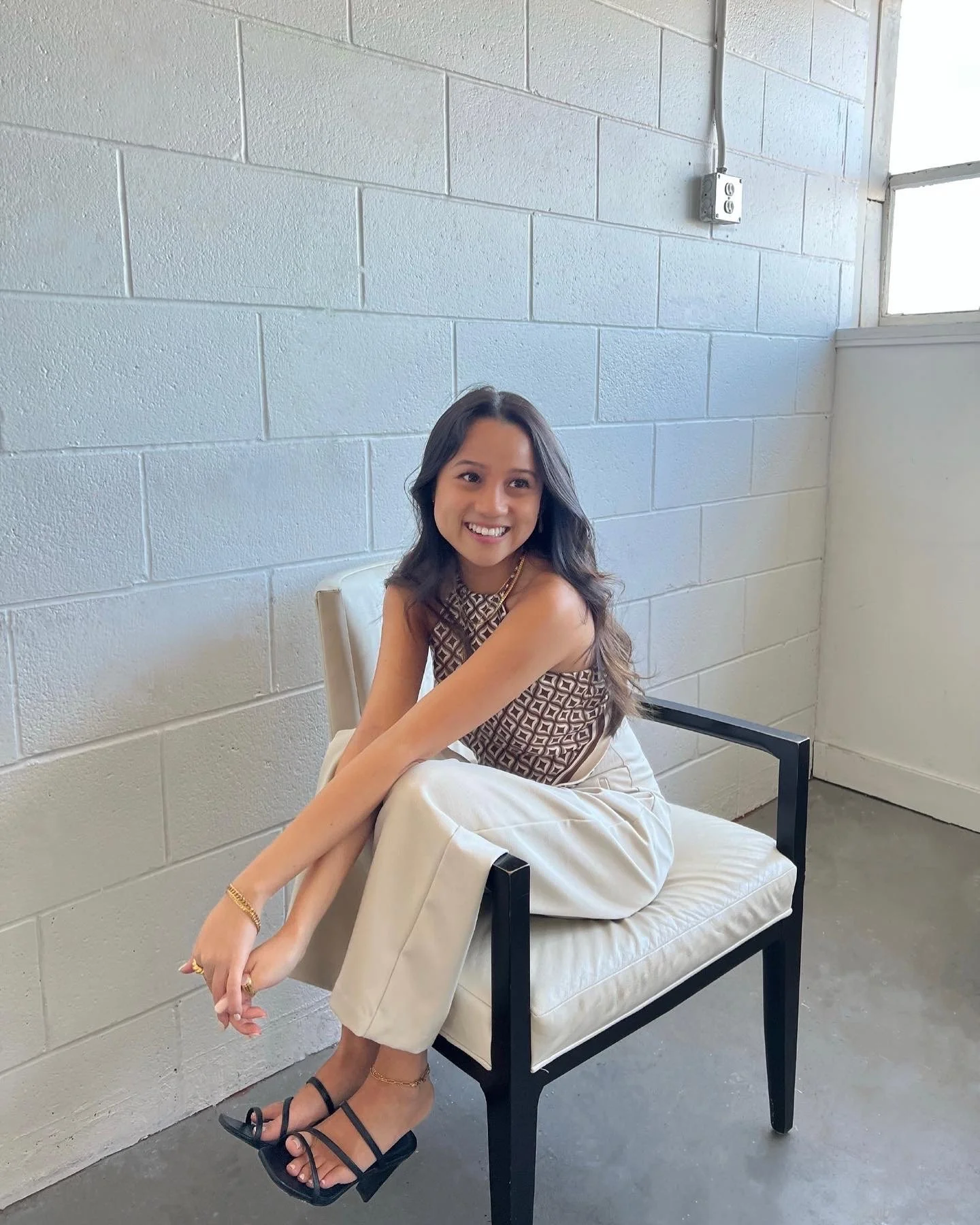 A young woman with long dark hair, wearing a patterned sleeveless top, cream-colored pants, and black high heels, sitting on a white chair with black frame, smiling in a minimalist room with white brick wall and window.