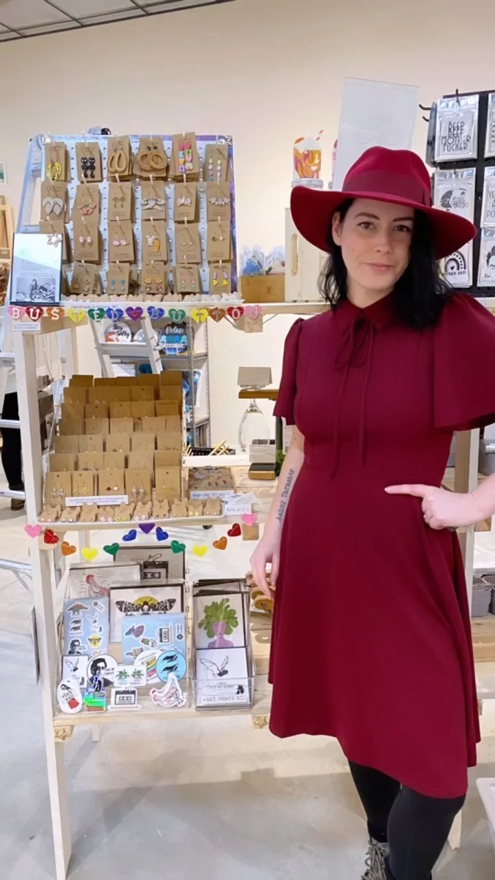 Woman in a red dress and matching wide-brim hat standing next to a display of jewelry and accessories at a craft fair or market.