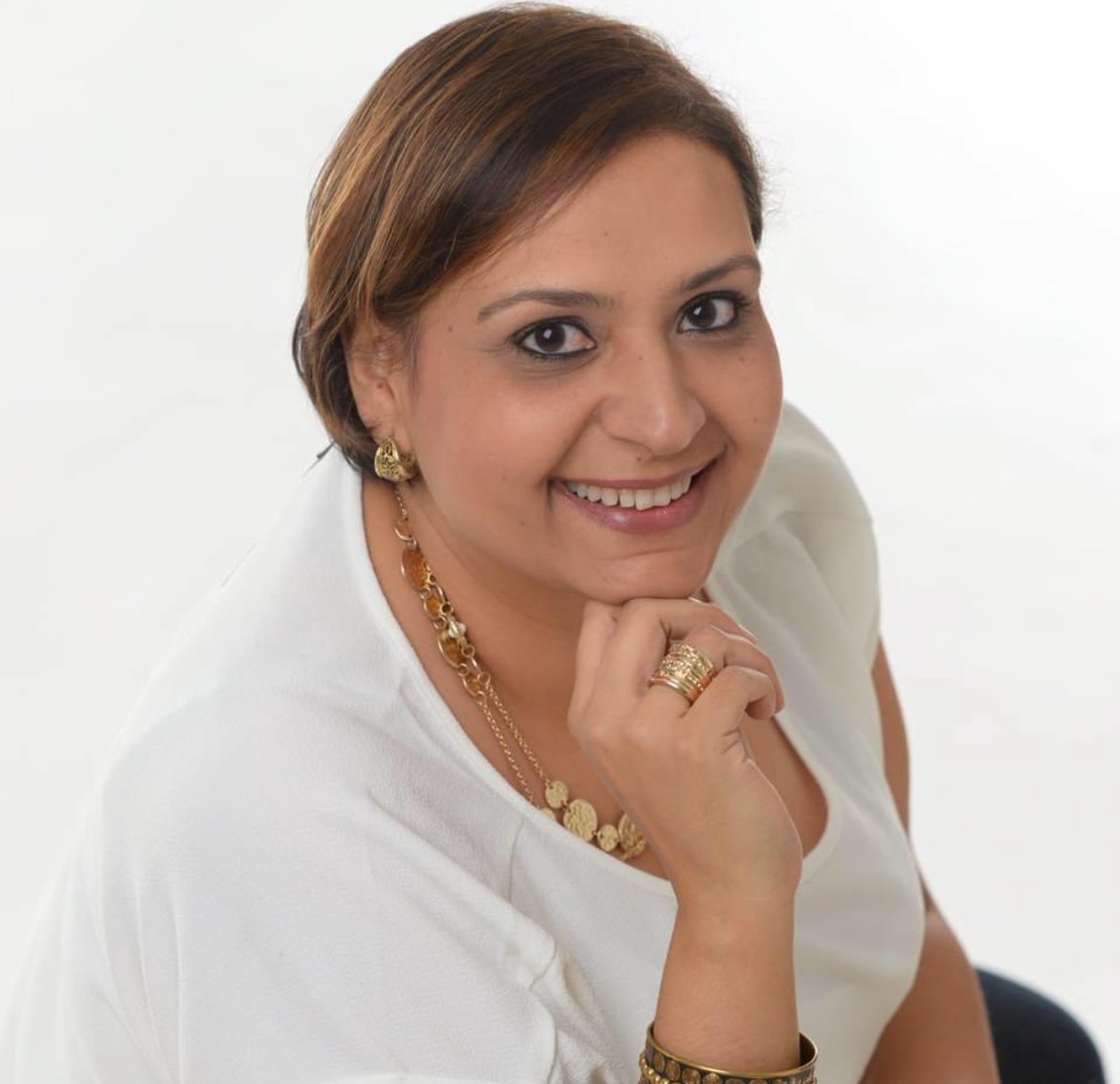 Close-up of a smiling woman with short brown hair, wearing gold jewelry and a white top, against a plain white background.