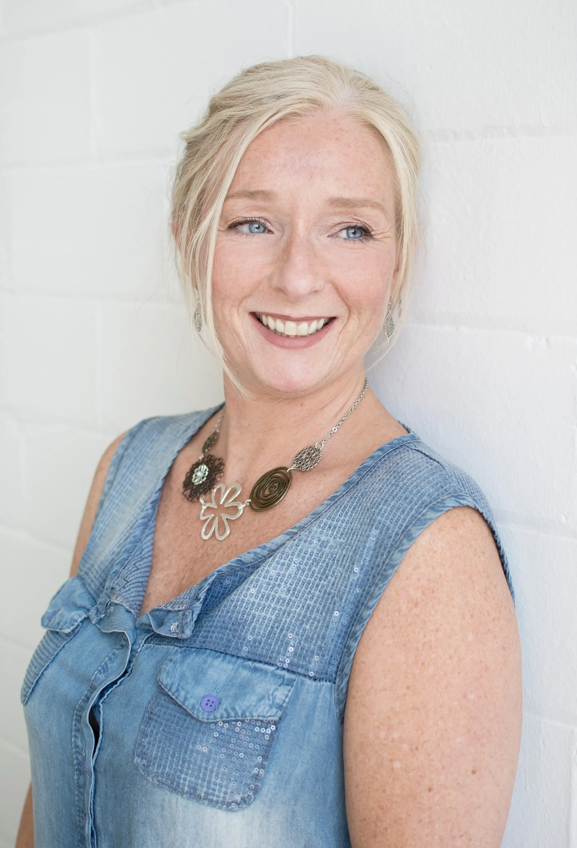 A smiling woman with blonde hair and blue eyes leaning against a white brick wall, wearing a sleeveless denim top and a large floral necklace.