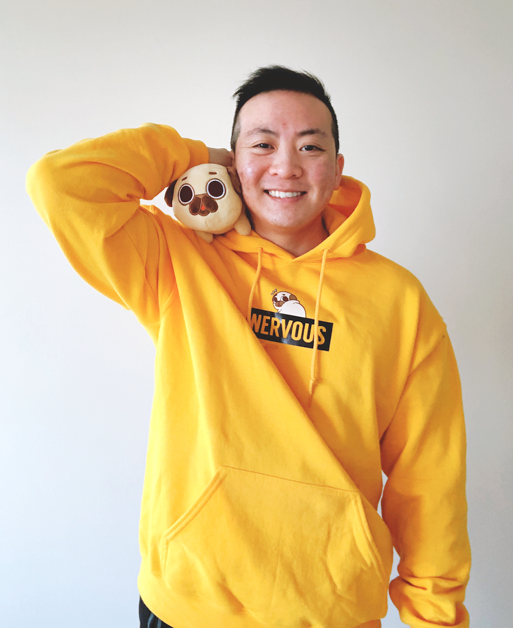 A young man with short black hair and a bright smile posing against a plain white wall. He's wearing a yellow hoodie with the word "NERVOUS" printed on it and is holding a plush toy of a pug dog on his shoulder.