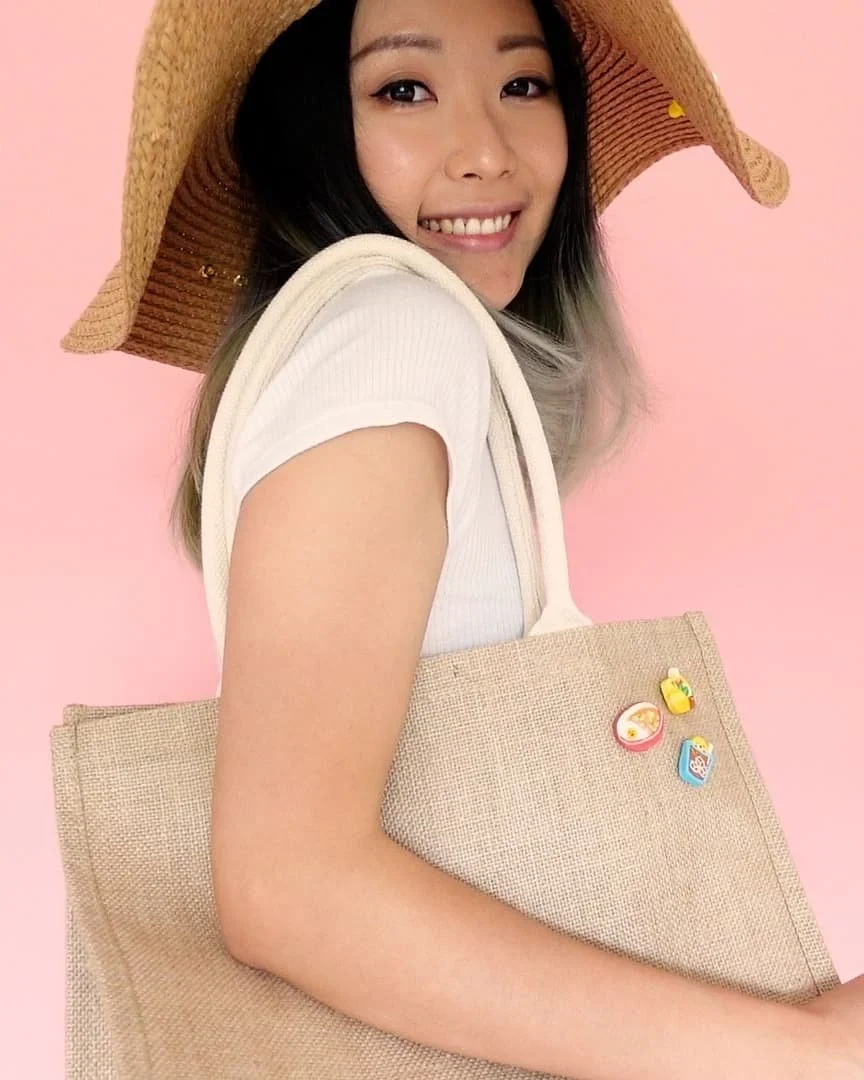 A young woman wearing a sunhat, smiling and looking at the camera, holding a beige tote bag with small colorful patches, standing against a pink background.