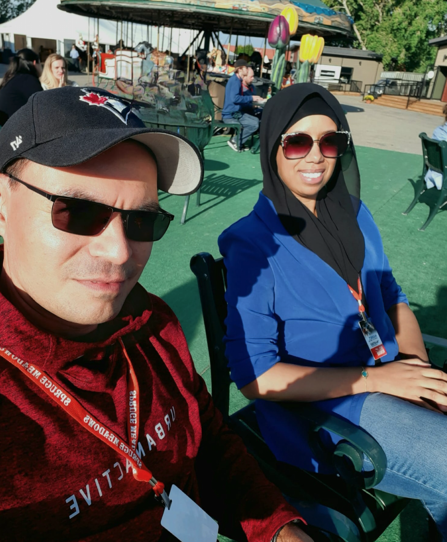 Two people sitting outdoors on green chairs, taking a selfie. The man on the left wears sunglasses, a black cap, and a red hoodie, while the woman on the right wears sunglasses, a black headscarf, and a blue jacket. In the background, there are people sitting at tables, inflatable yellow tulips, a carousel, and trees.