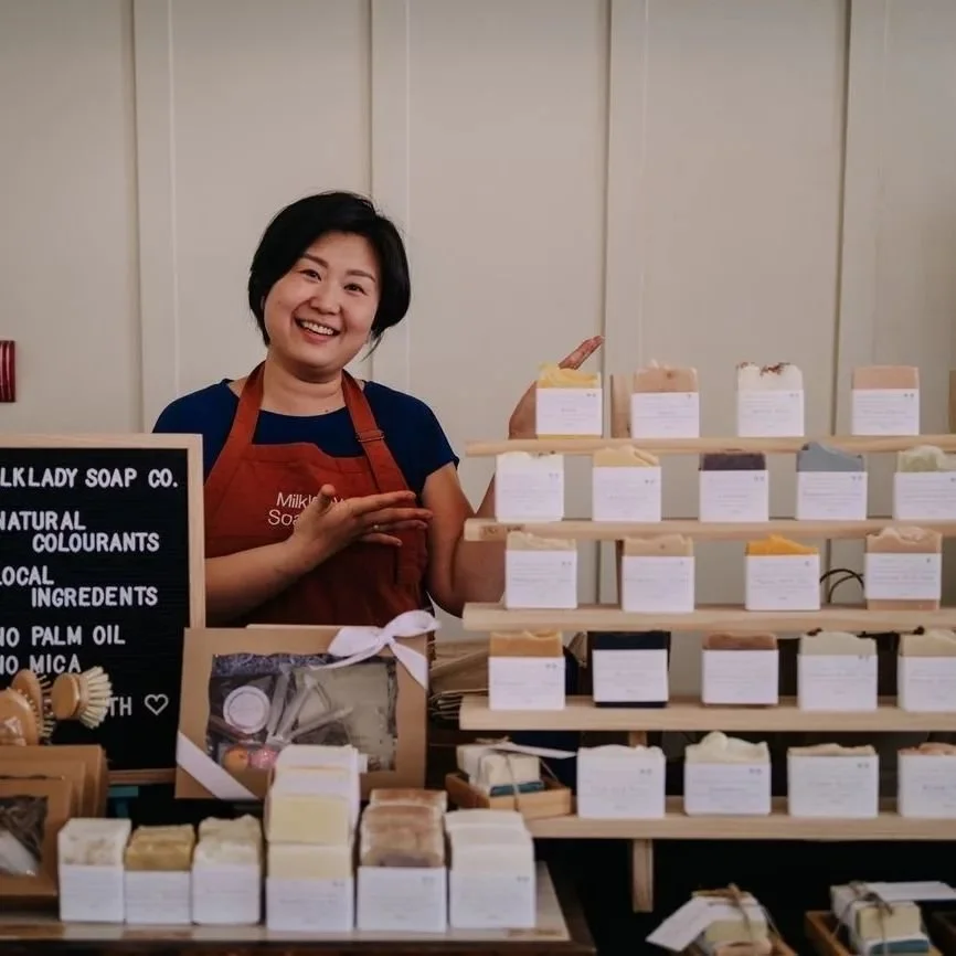 A woman smiling at a soap booth with various handmade soaps displayed on shelves and counters.