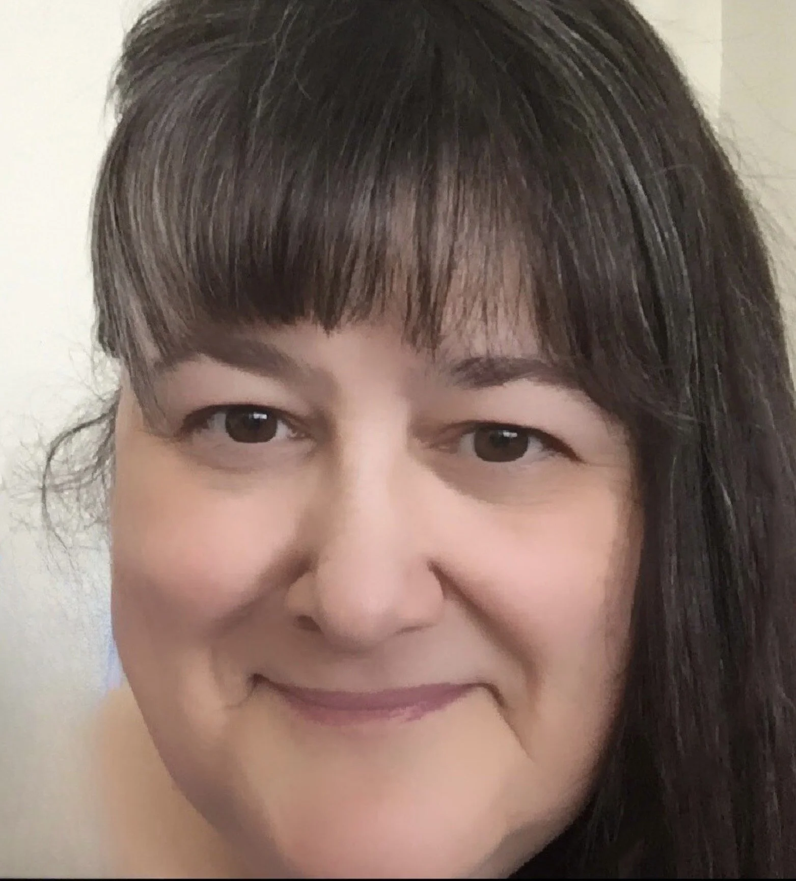 A close-up photograph of a woman with dark brown hair with bangs, smiling softly, with light makeup and fair skin.