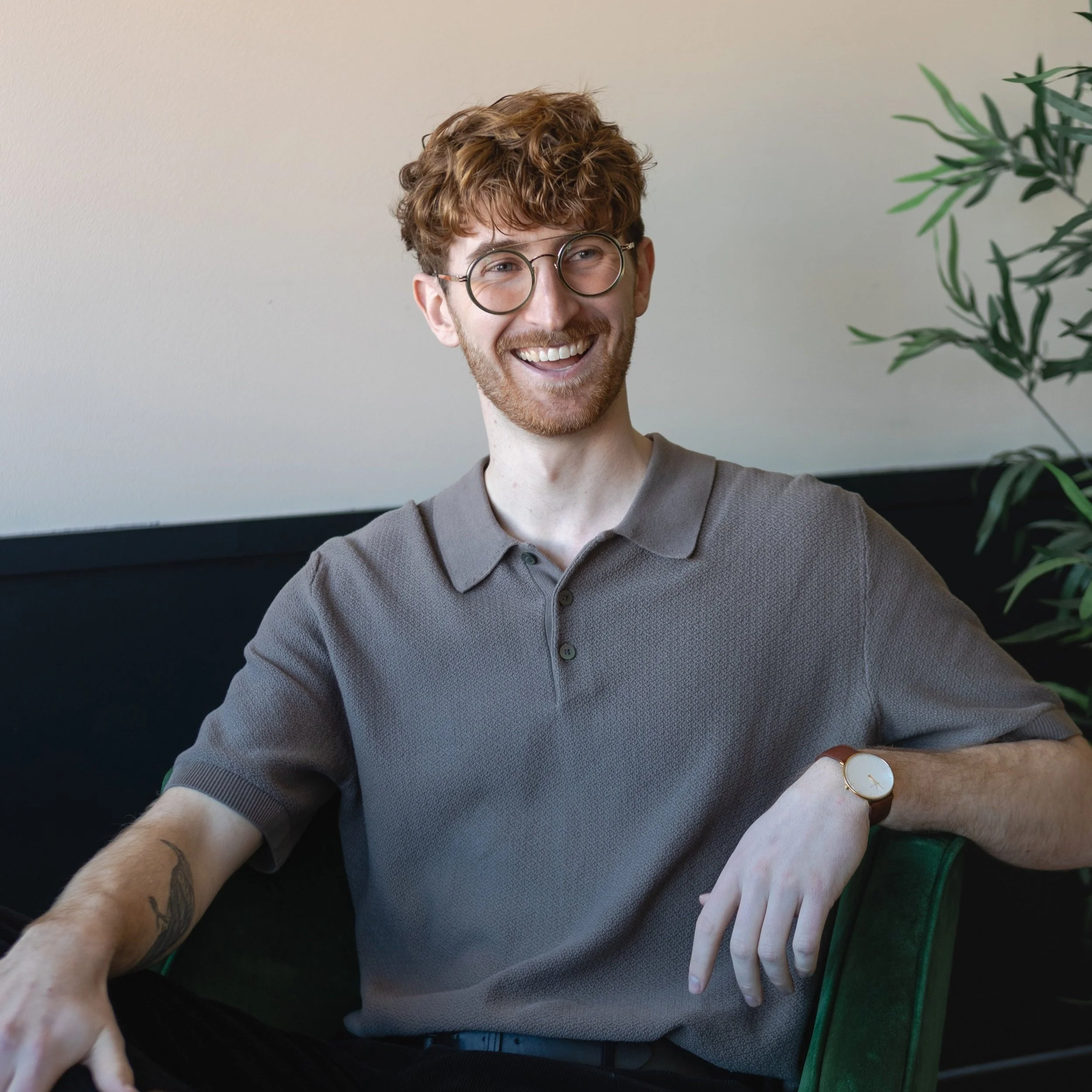 A smiling man with curly red hair, glasses, a beard, wearing a gray polo shirt, a watch, and arm tattoos, sitting in a green chair next to a plant.