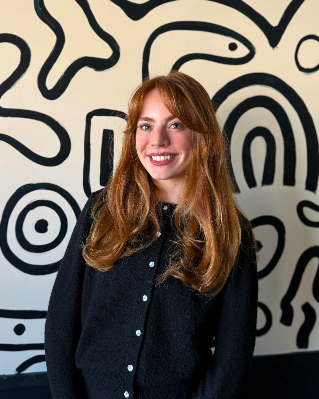 A smiling woman with long, wavy red hair and wearing a black sweater, standing in front of a white wall with abstract black lines and shapes.