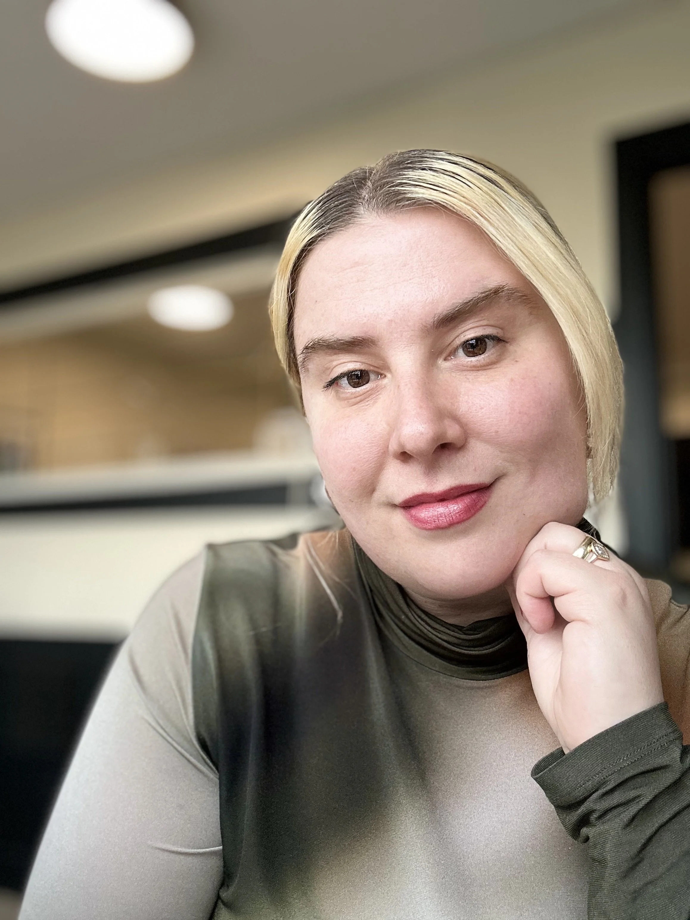 Close-up of a woman with blonde hair, wearing a black and beige top, resting her chin on her hand, smiling softly in an indoor setting with blurred background.