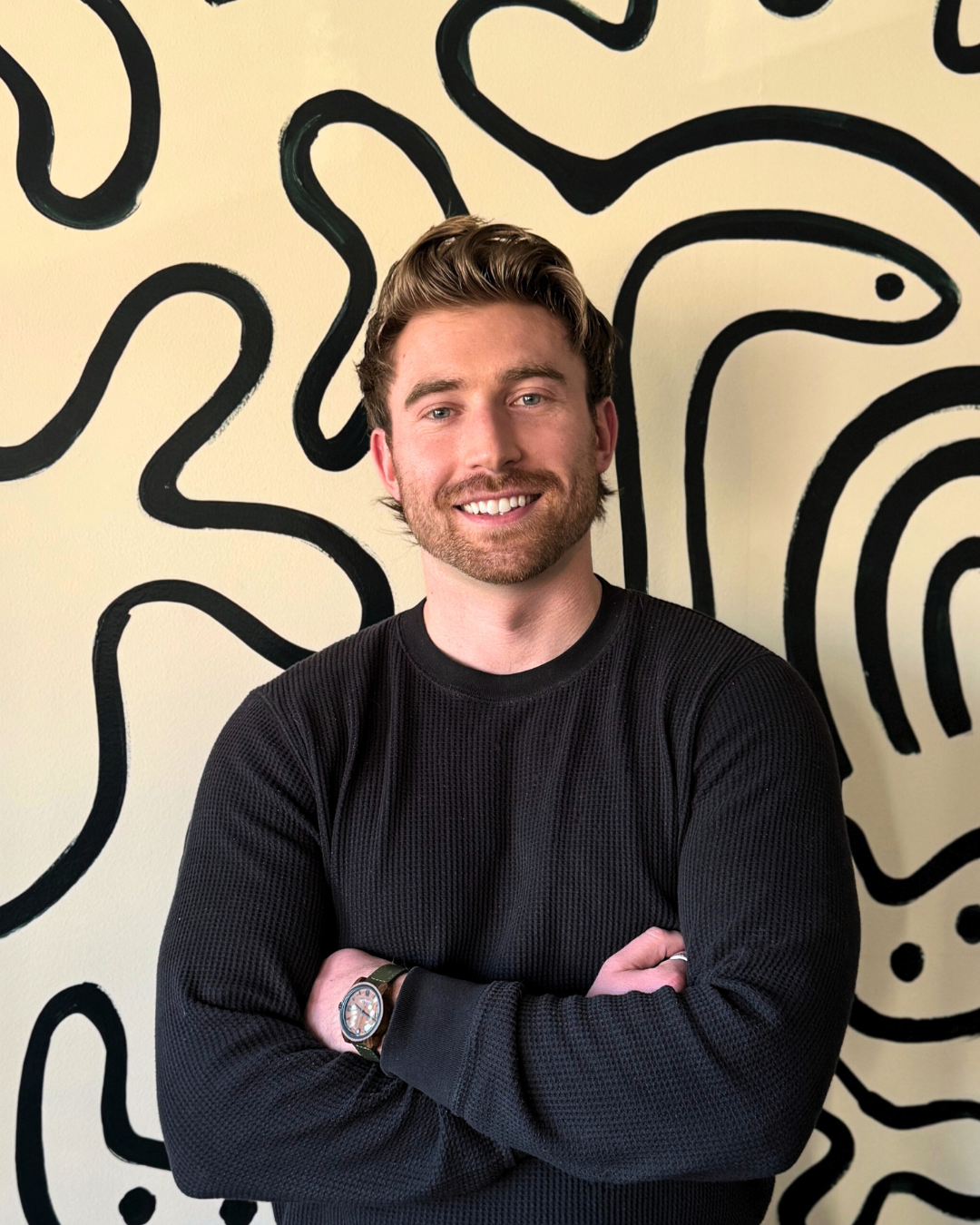 A man with brown hair and a beard, smiling with crossed arms, standing in front of a wall with black abstract line art.