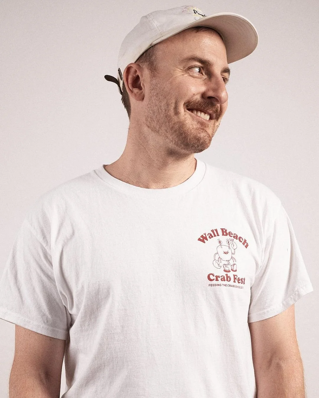 A smiling man wearing a white t-shirt with a crab graphic and text that reads 'Wall Beach Crab Fest,' and a white baseball cap against a plain light background.