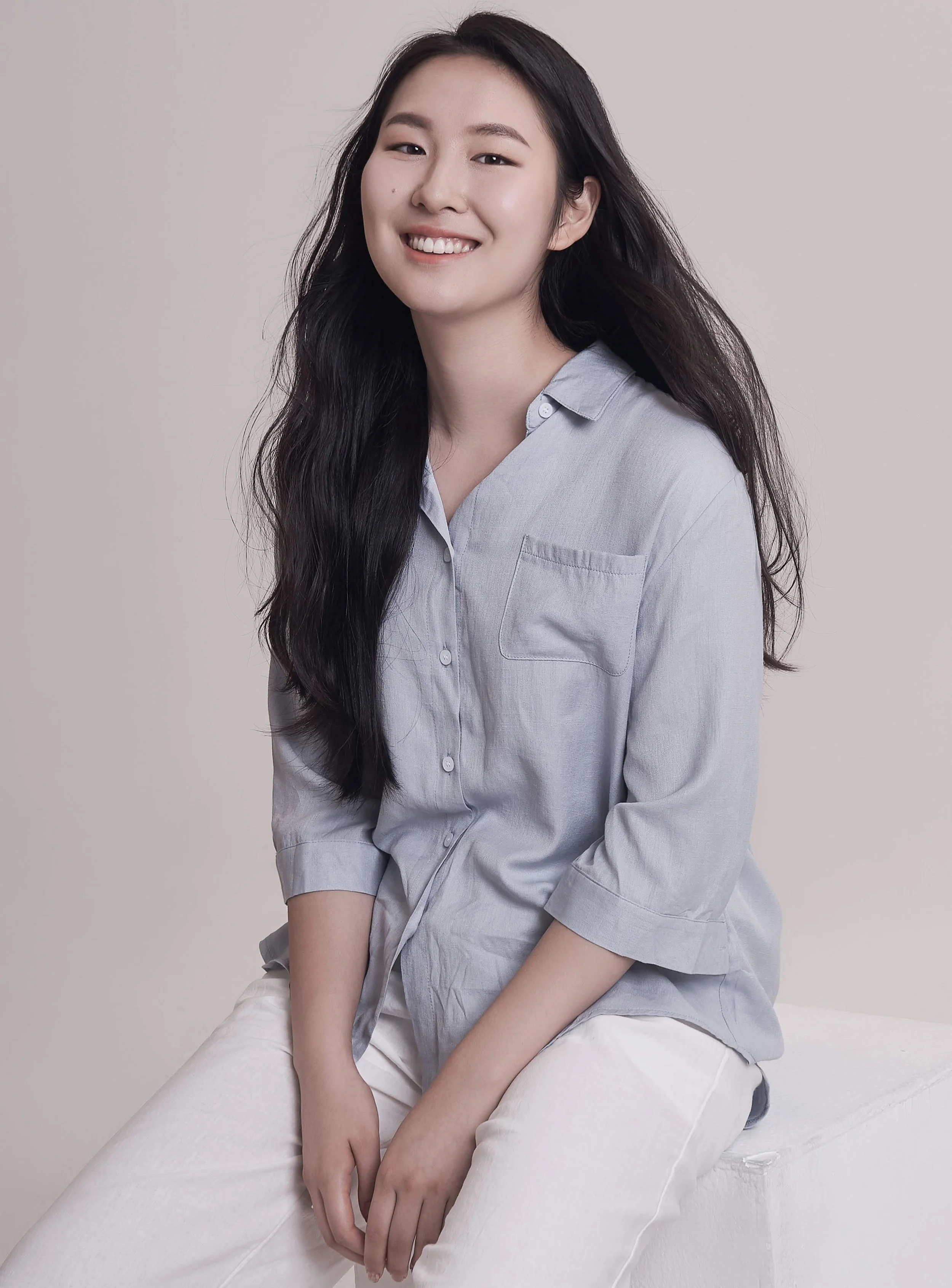 A young woman with long black hair smiling softly, seated on a white surface against a plain light-colored background, wearing a light gray button-up shirt and white pants.