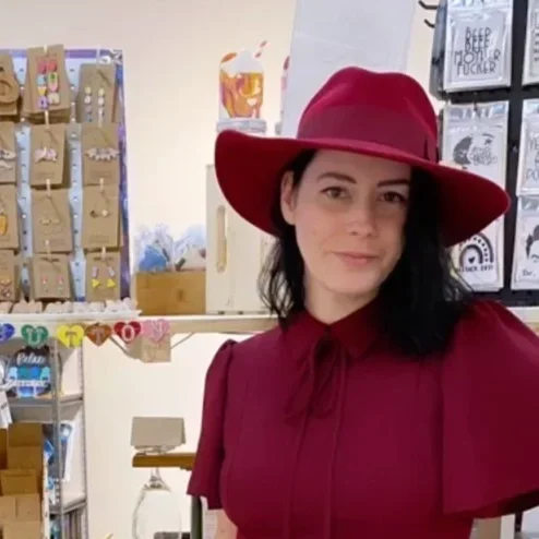 Woman in a red hat and dress standing in a store with shelves of snacks, a beverage, and store displays in the background.