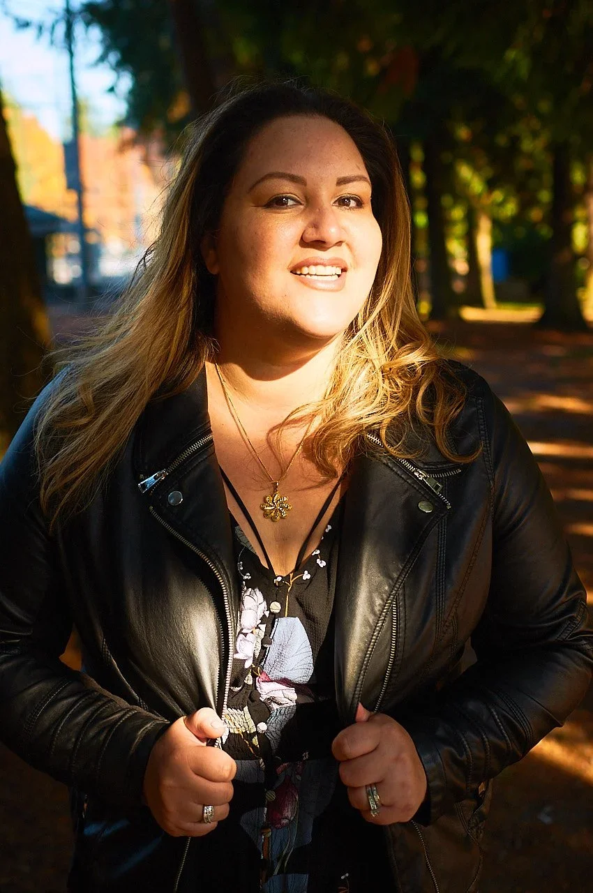 A woman with long wavy blonde hair smiling outdoors in a park with trees and sunlight in the background, wearing a black leather jacket and layered necklaces.