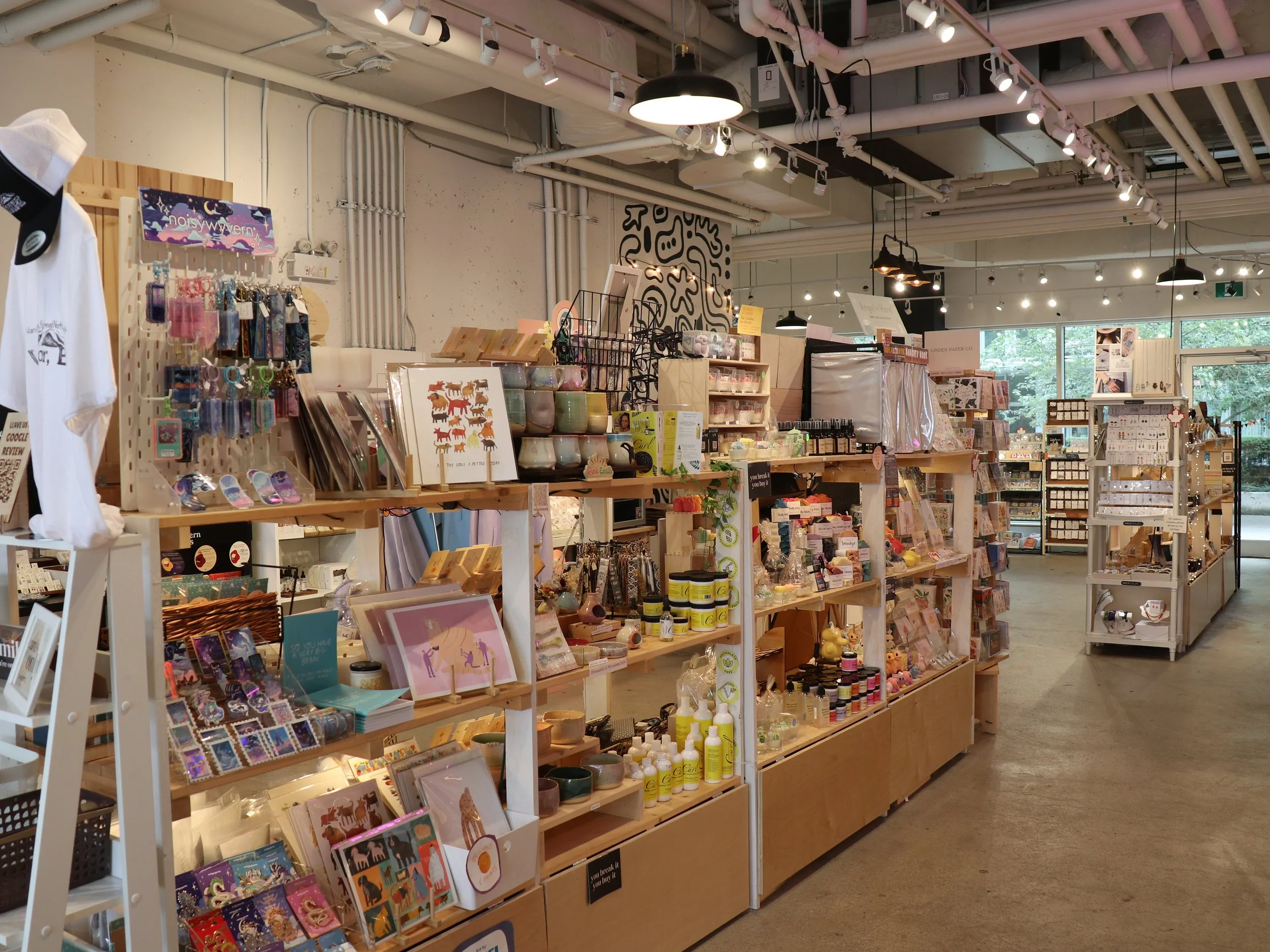 Interior of a retail store with wooden shelves displaying various colorful items including stationery, candles, artwork, and accessories. Large windows and industrial ceiling lights. The store appears bright and organized.