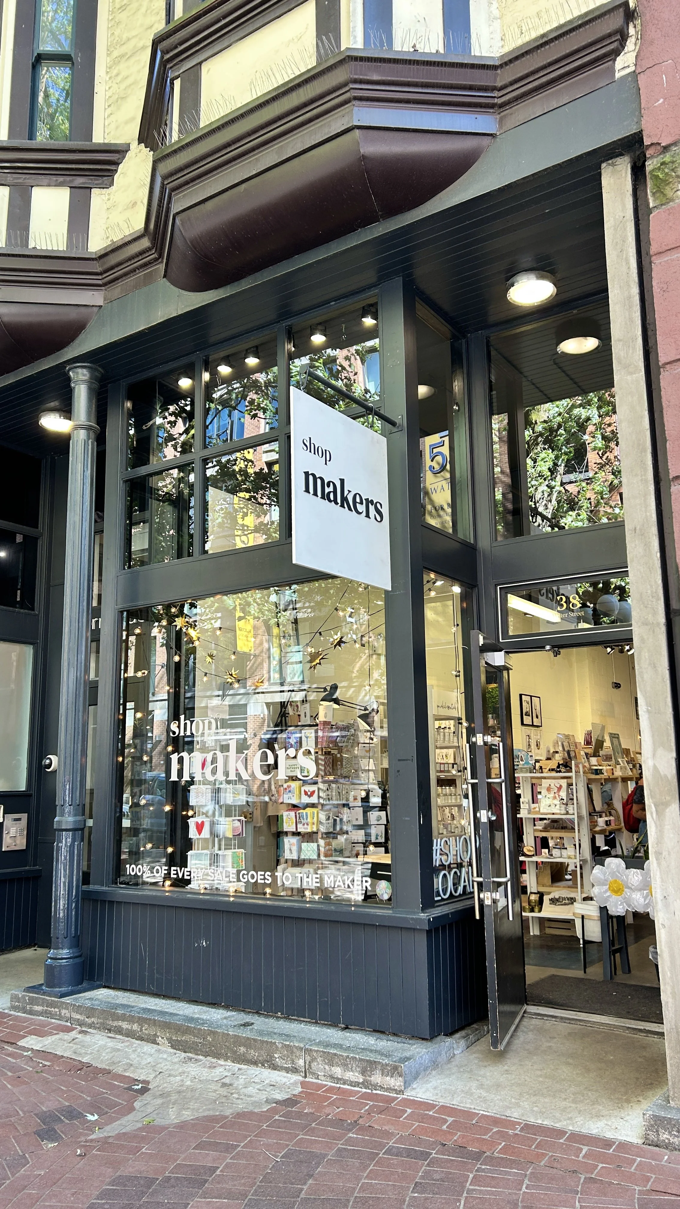 Exterior of a shop named 'shop makers' with large glass windows showing the interior, which has shelves with products and artwork; the shop has a black frame, and there's a sign with the shop's name hanging outside.