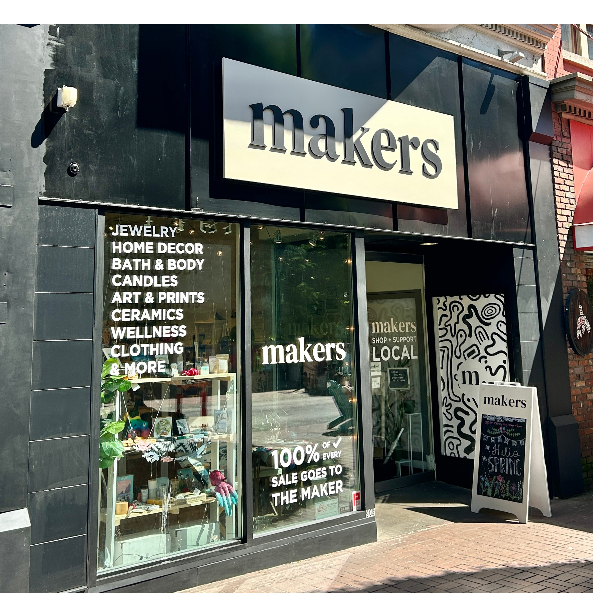 Exterior view of a store titled 'makers' with glass windows displaying various products, and a sign advertising categories like jewelry, home decor, bath & body, candles, art & prints, ceramics, wellness, and clothing.