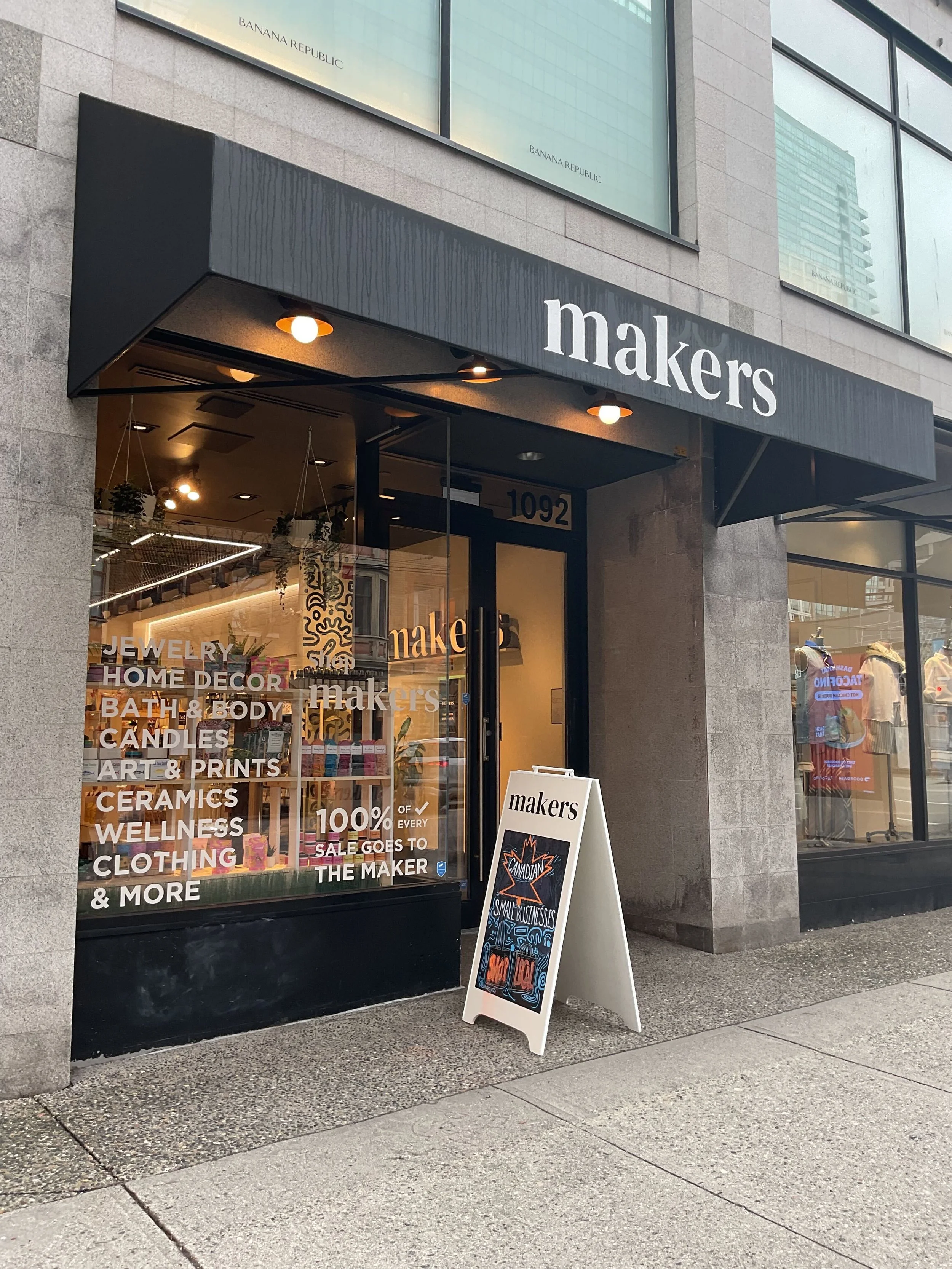 Outside view of a store called Makers, with a glass door and window display showcasing various products and signs indicating items like jewelry, home decor, bath and body, candles, art prints, ceramics, wellness, and clothing. A sidewalk and digital sign advertising a small business event are also visible.