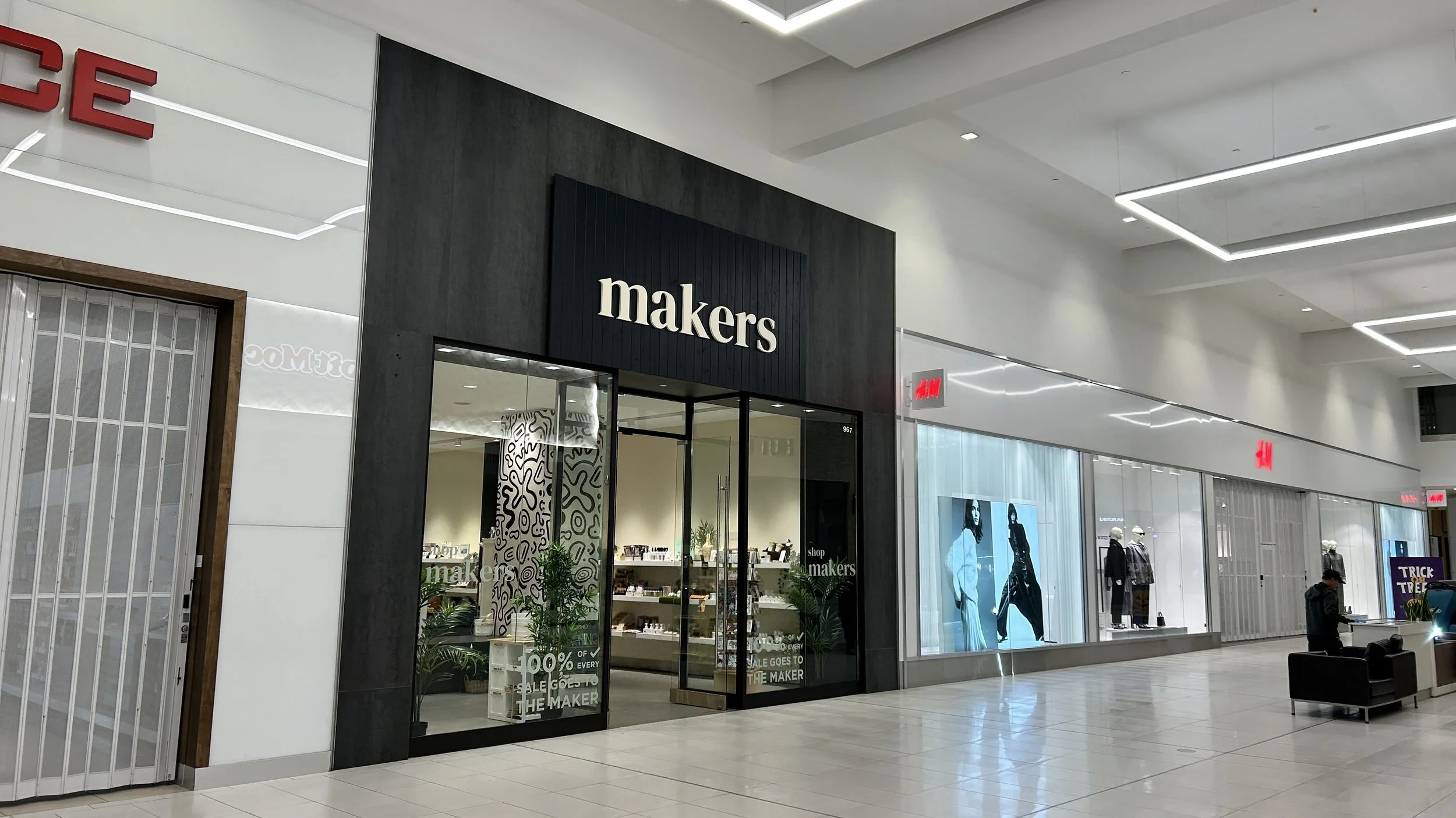 Inside a shopping mall, the image shows the storefront of a store called 'makers' with a glass door and windows displaying merchandise. Adjacent to it are other retail stores, including an H&M store with mannequins and mannequins, and digital screens displaying fashion models. There is a seating area with a person sitting nearby, and the mall has a modern ceiling with recessed lighting.