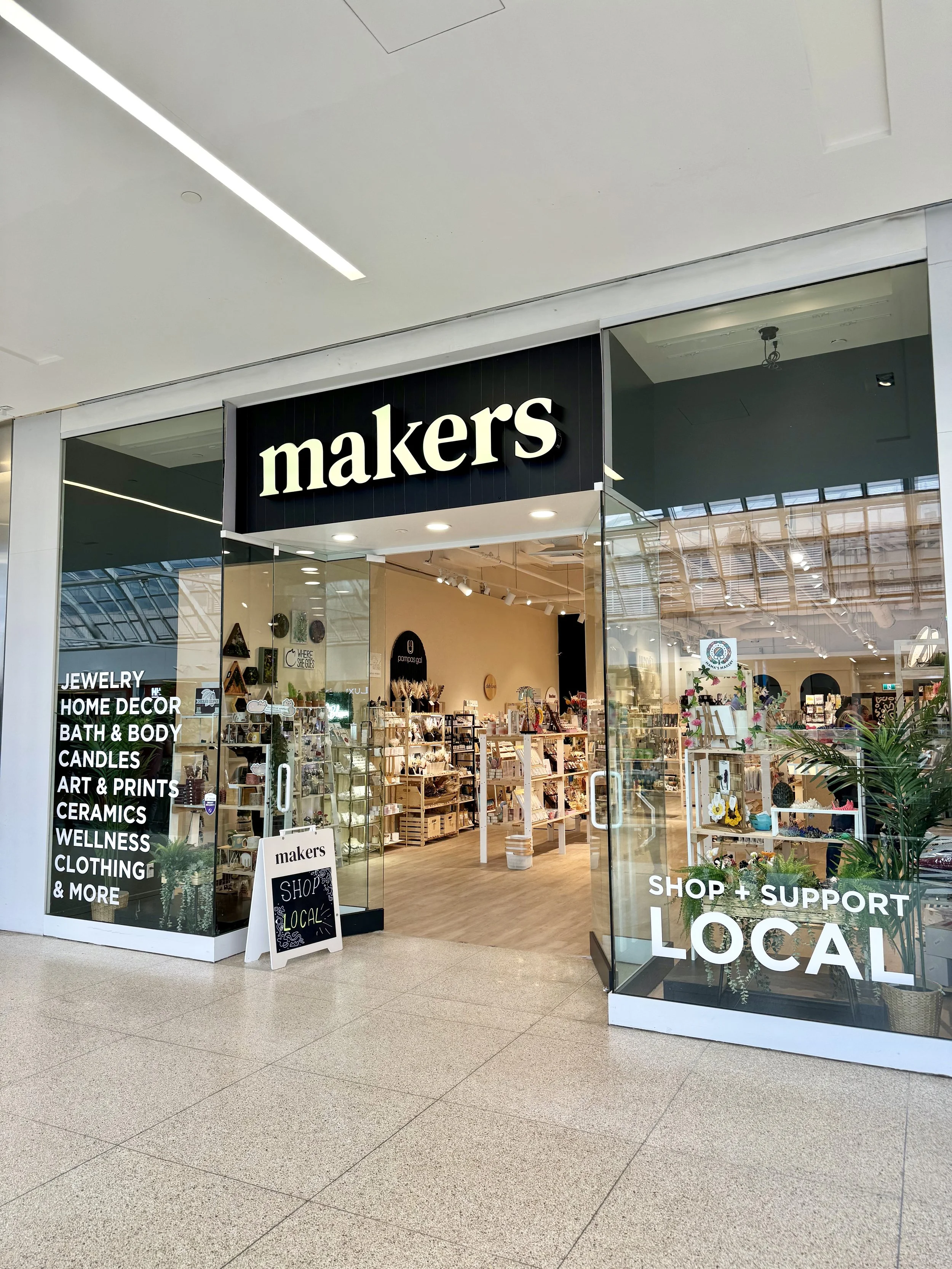 Entrance of a shop named 'makers' with a glass storefront showcasing various artisan products inside. Sign on the window lists categories like jewelry, home decor, bath & body, candles, art & prints, ceramics, wellness, clothing, and more. A chalkboard sign outside promotes shopping local.