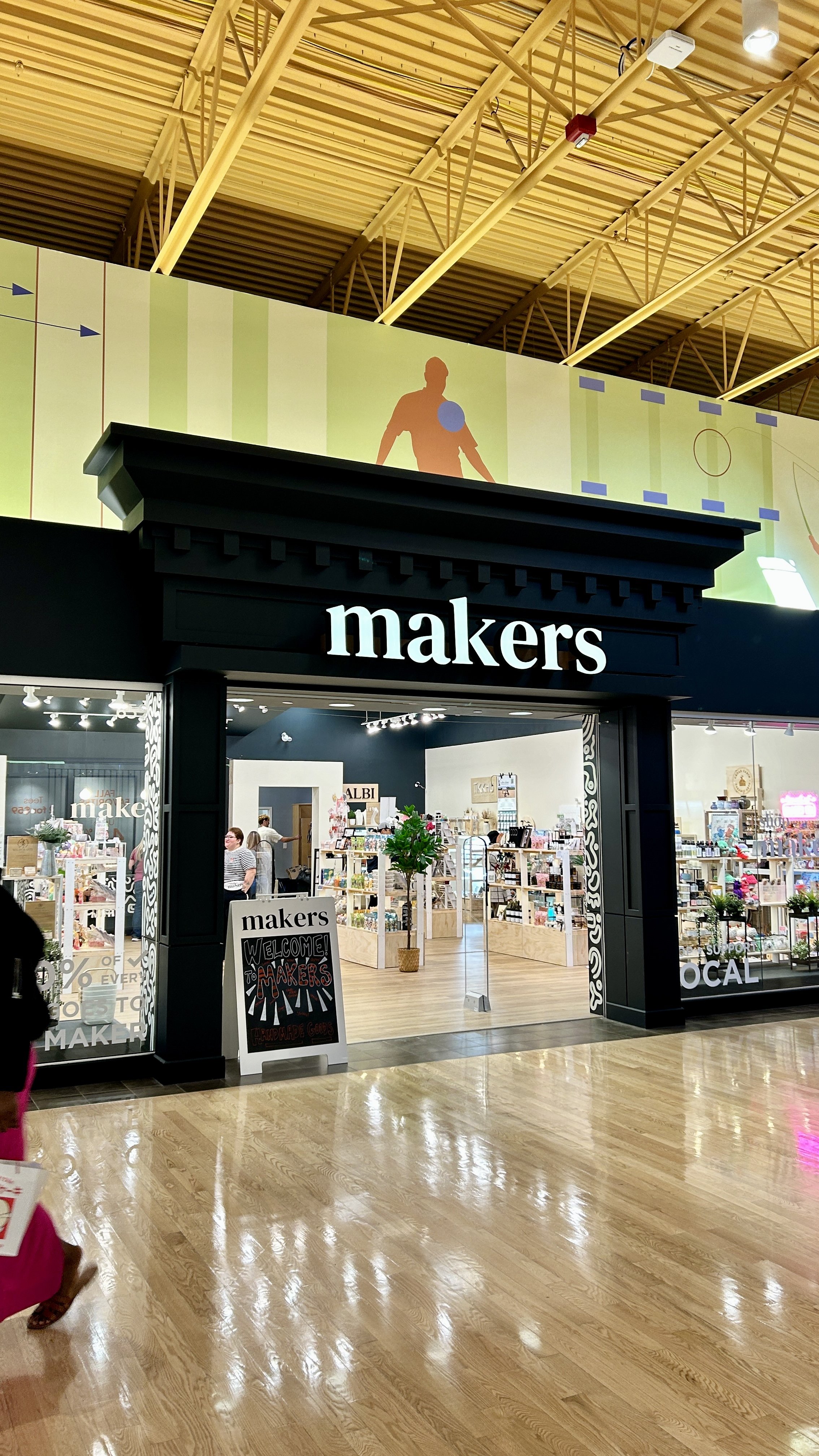 Entrance to a store called 'makers' with a black facade and a sign above, inside are various craft and handmade items displayed on shelves, with customers shopping.
