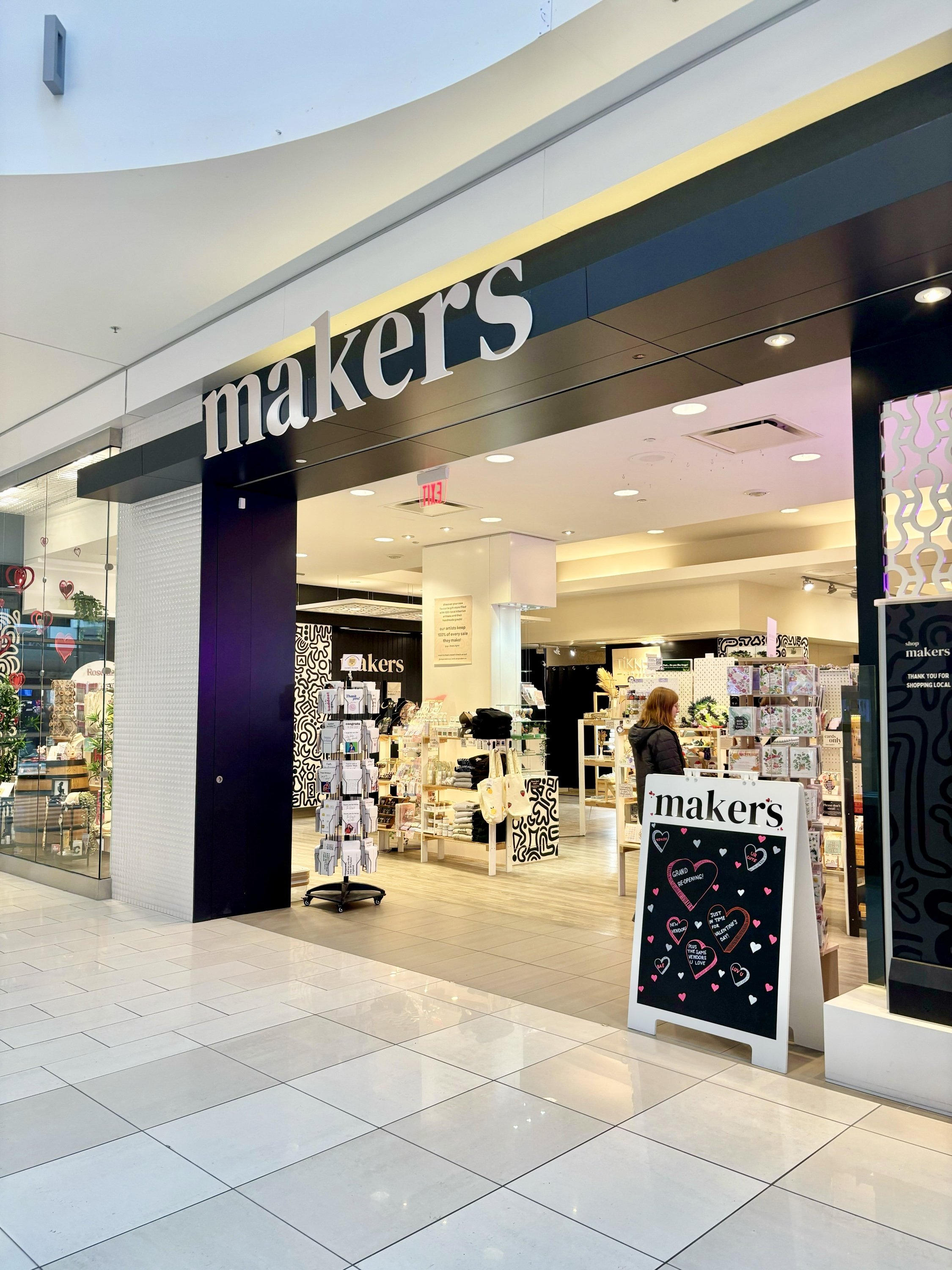 Entrance to a store called Makers, with a black sign and white lettering. Inside, various display shelves with products, and a woman browsing. A chalkboard sign stands outside.