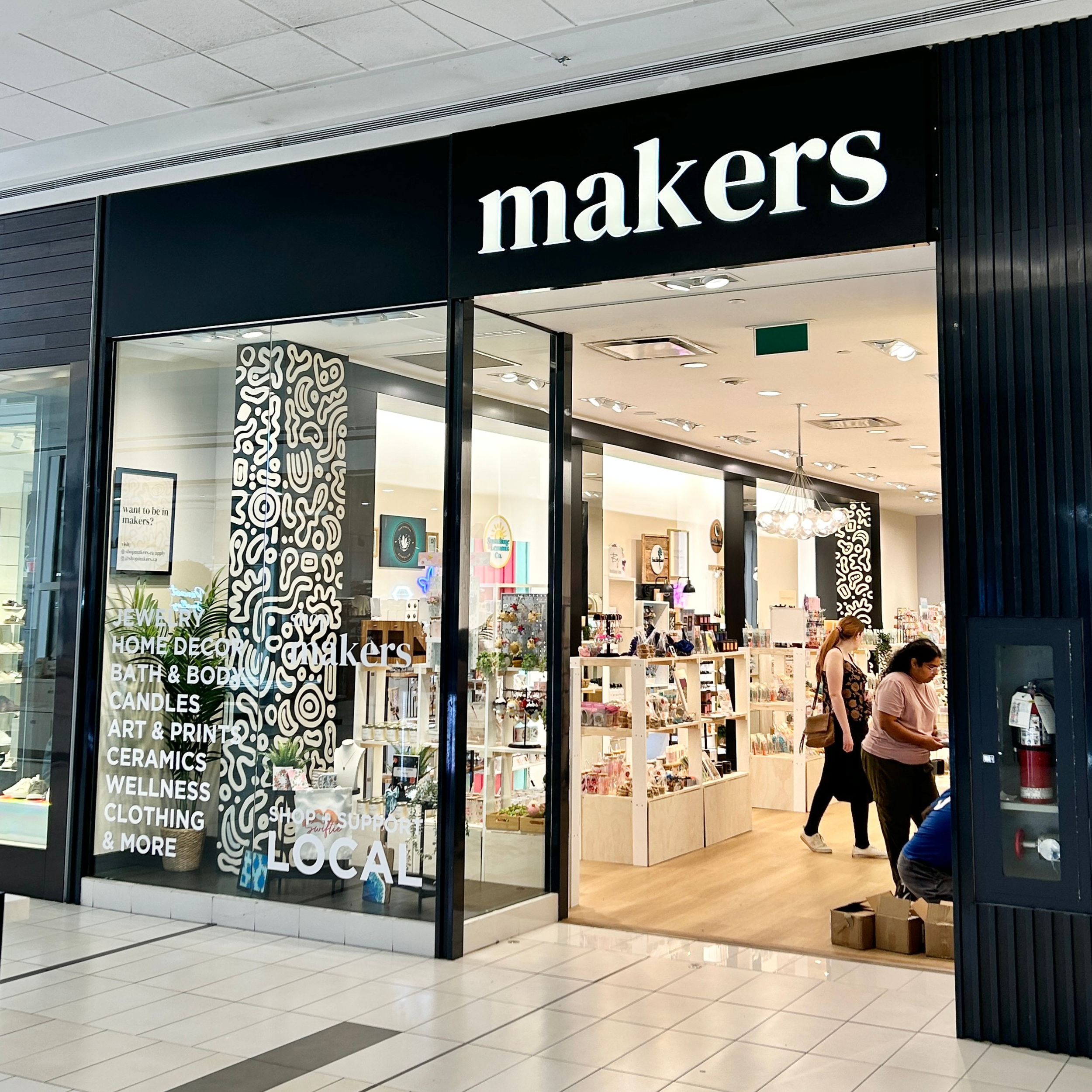 View of the storefront for Makers, a retail store selling jewelry, home decor, bath and body products, candles, art and prints, ceramics, wellness items, clothing, and more, with shoppers inside browsing.