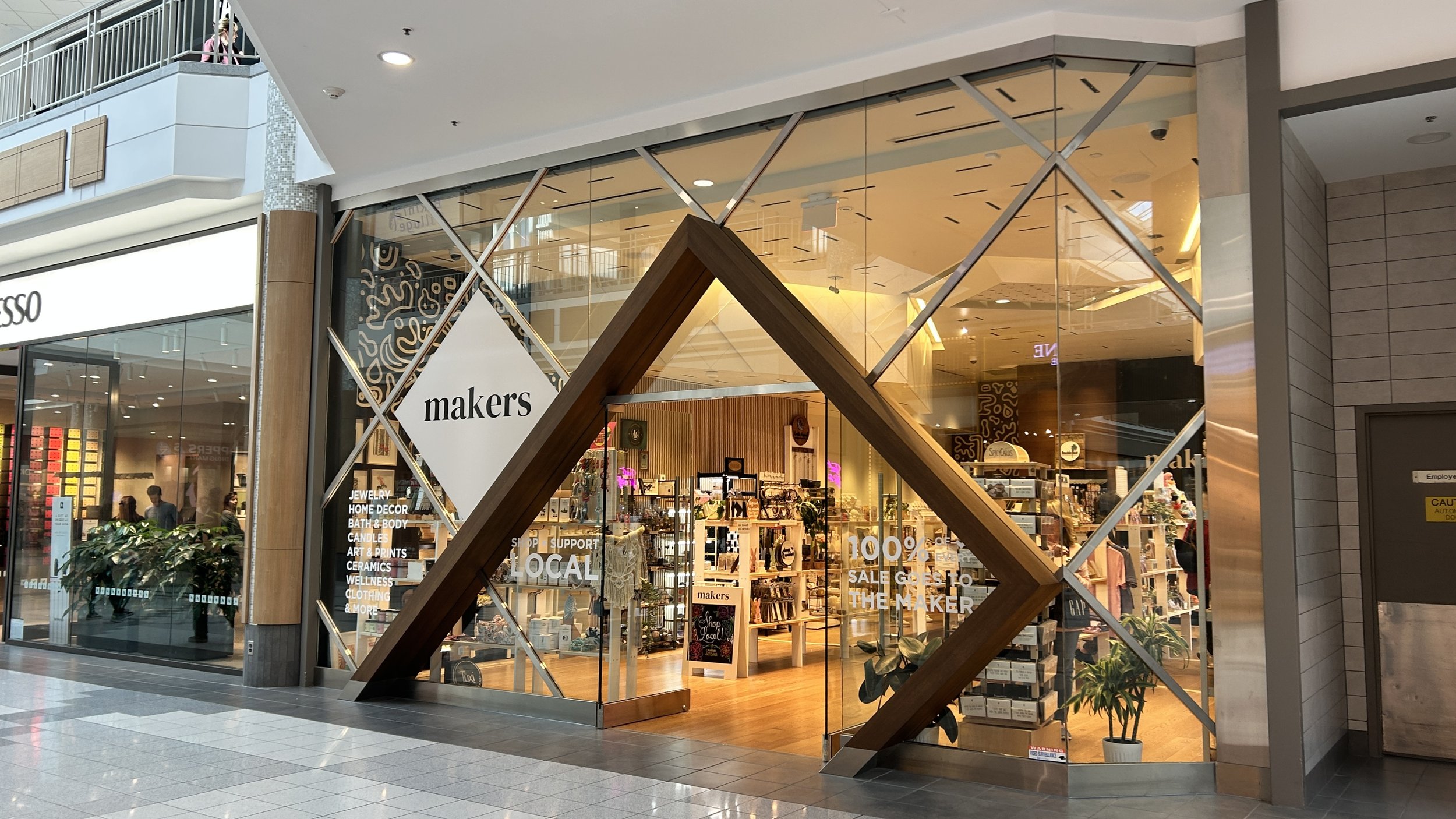 The storefront of a shop called "makers" in a shopping mall, with large glass windows and wooden accents. Inside, various products are displayed on shelves, and there is signage indicating the shop supports local makers and offers a sale. The mall exterior shows a tiled floor and neighboring stores.