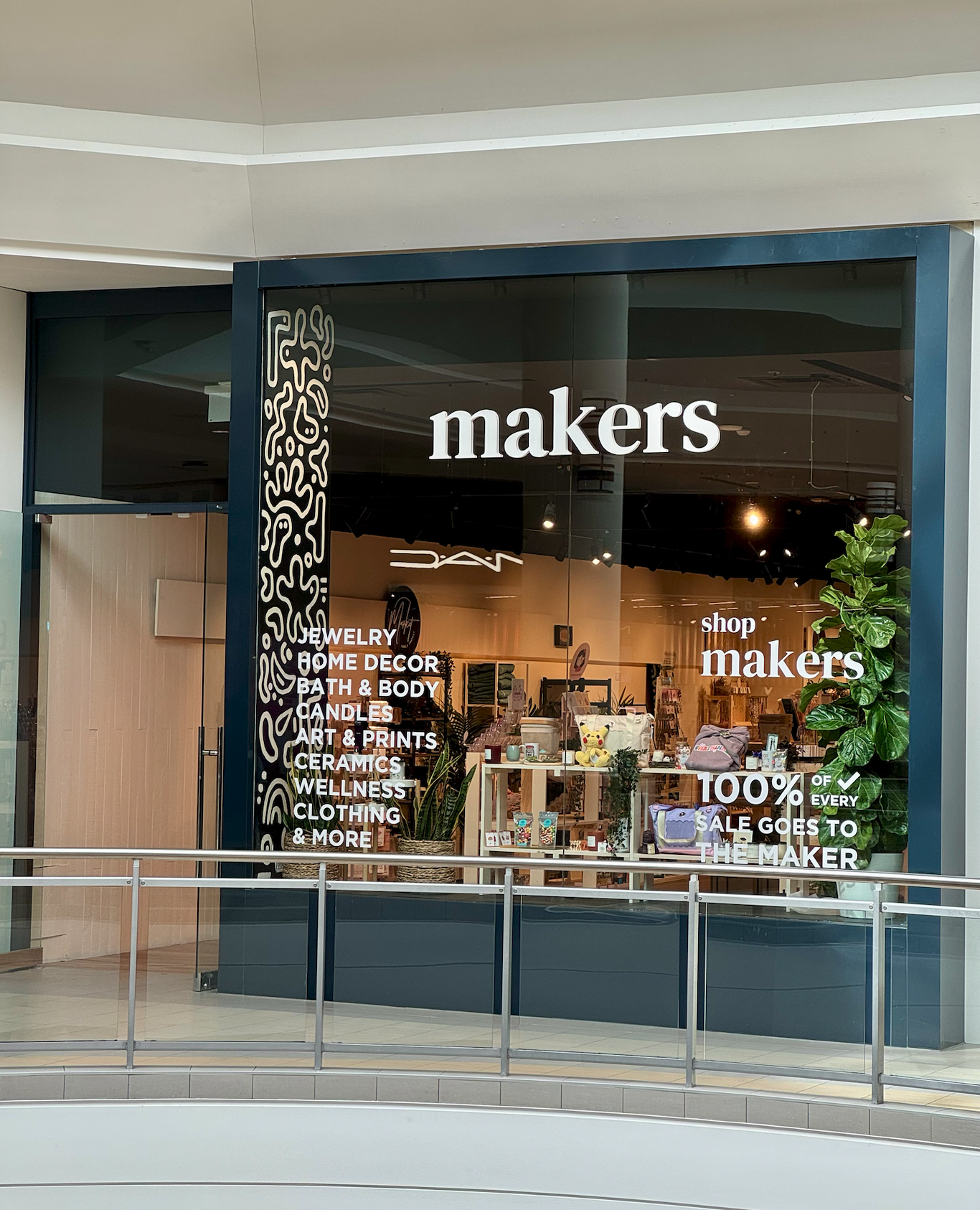Storefront with the name 'makers' and list of products like jewelry, home decor, bath & body, candles, art & prints, ceramics, wellness, clothing, and more, with a large green plant outside and signage indicating 100% of sales go to the maker.