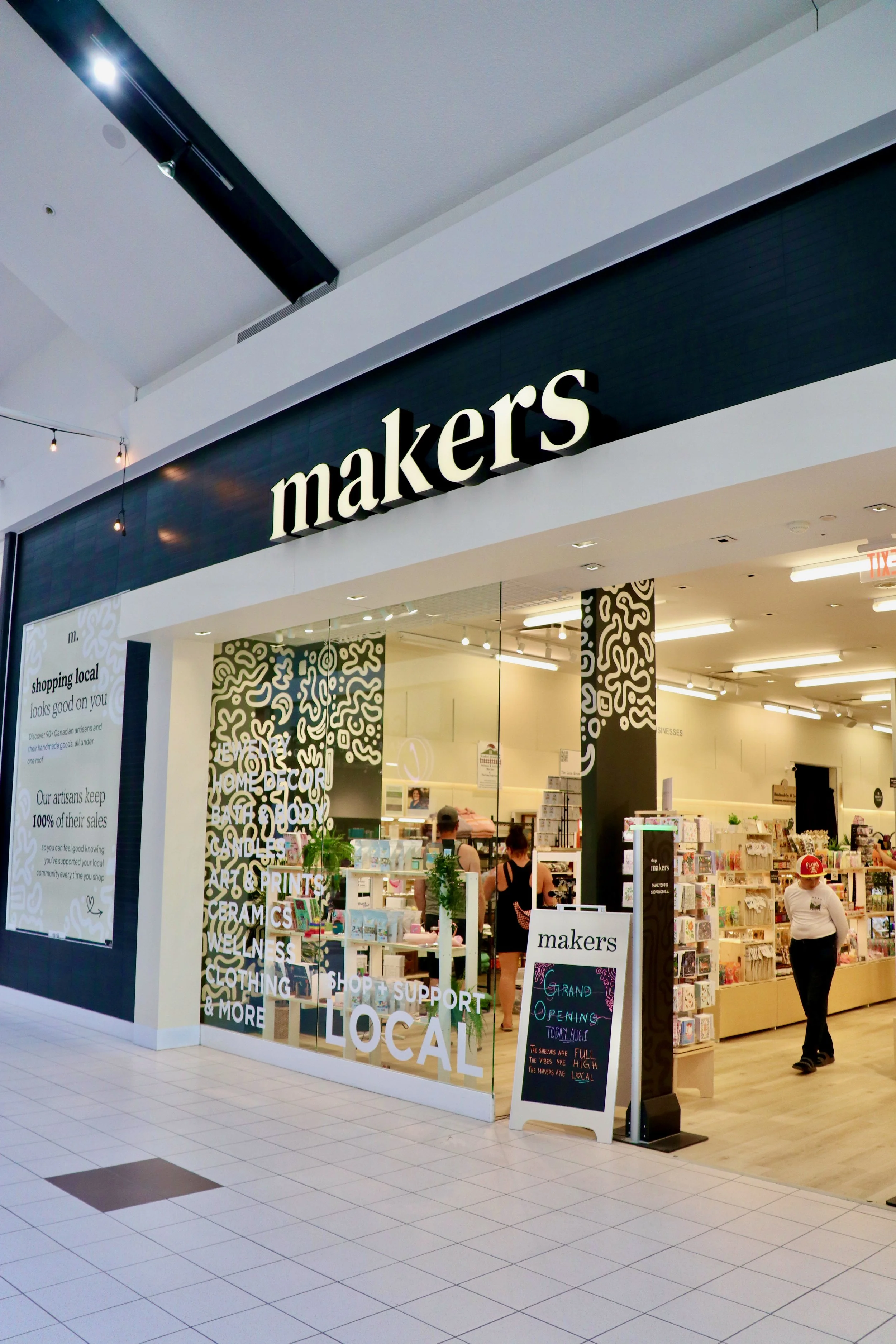 Exterior view of a store named 'makers' inside a shopping mall, with people shopping and a sign announcing a grand opening.