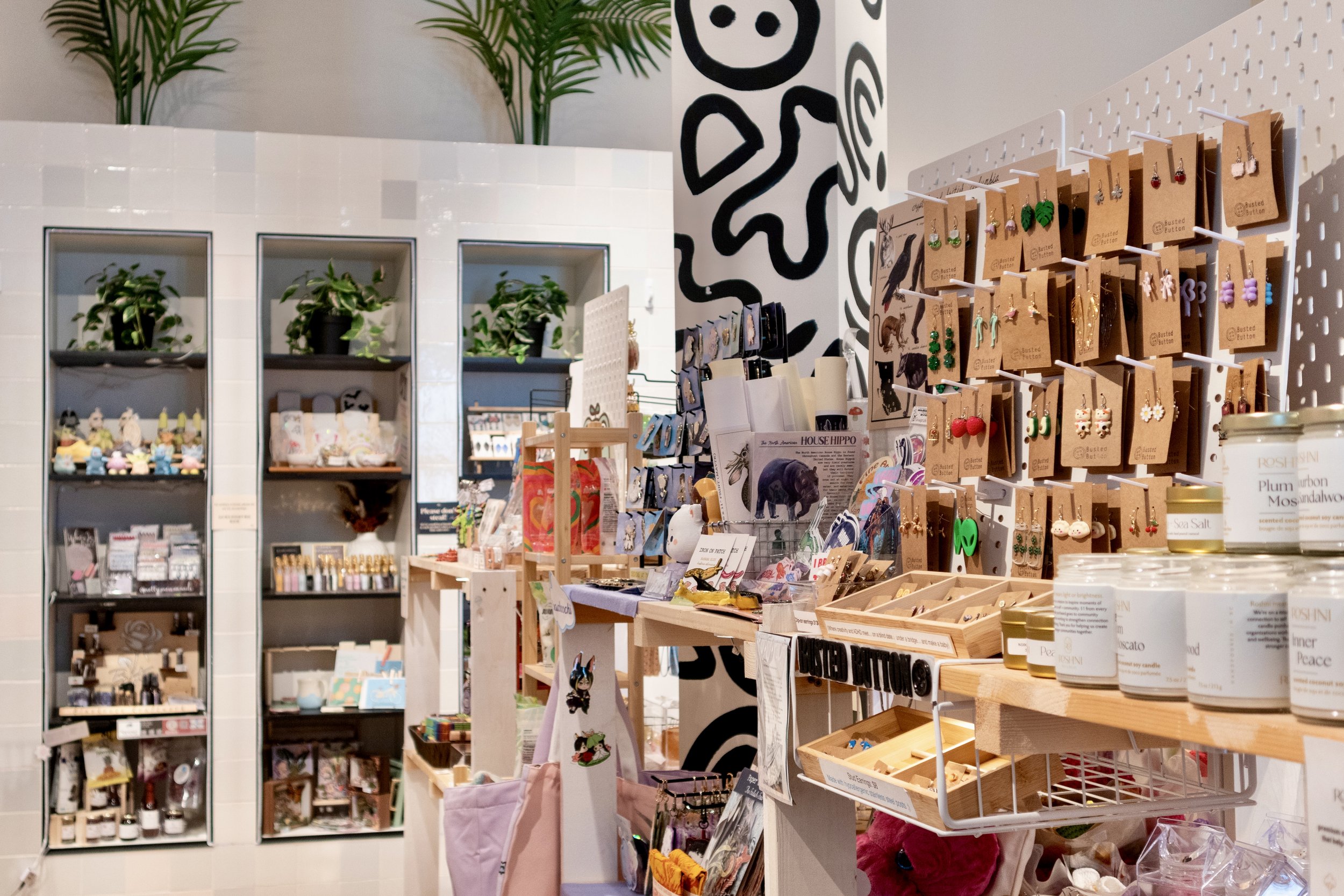 Display of various jewelry, accessories, candles, and small decorative items in a boutique store with potted plants and black and white abstract wall art.