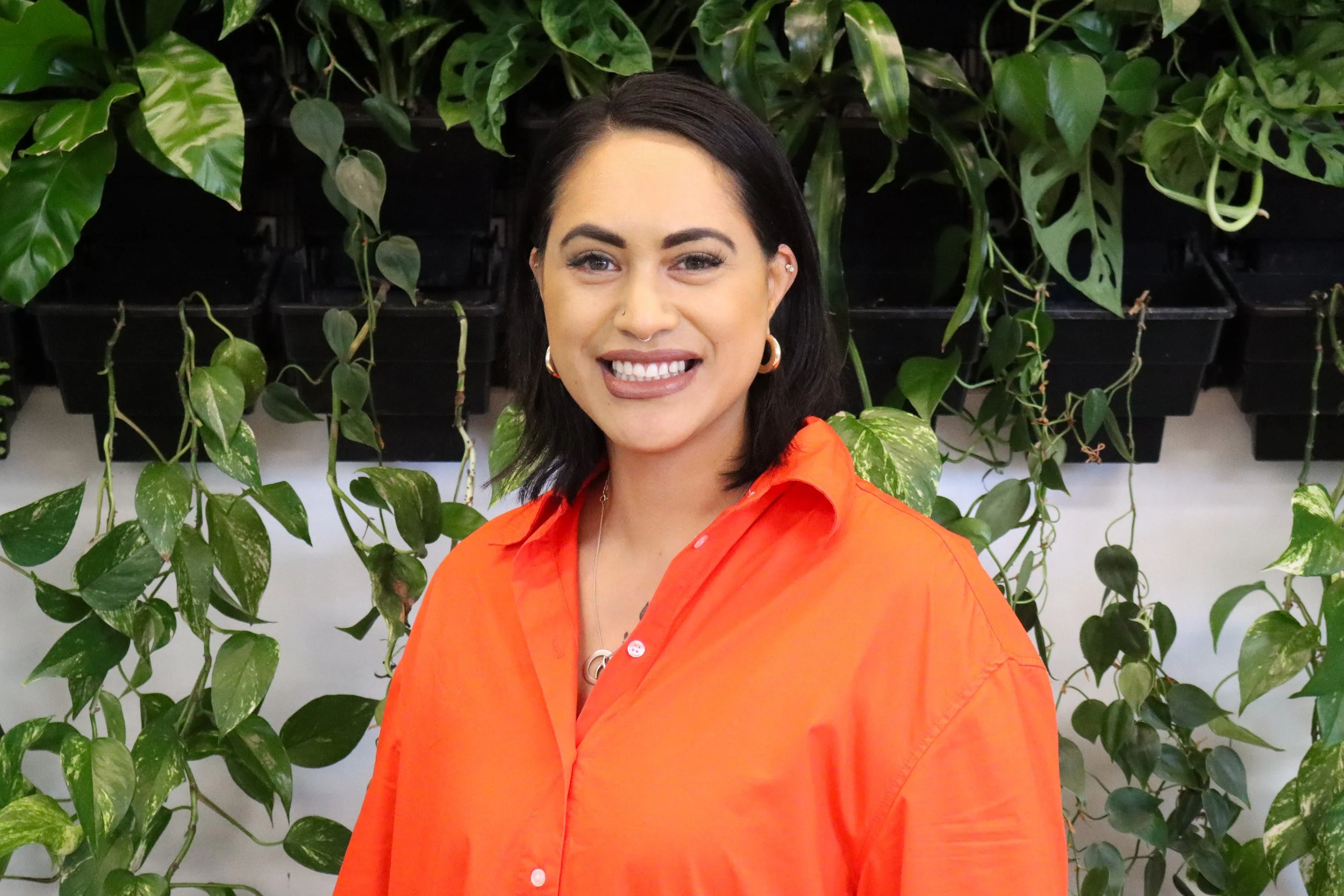 A woman with dark hair wearing an orange shirt, smiling in front of a wall of green leafy plants.