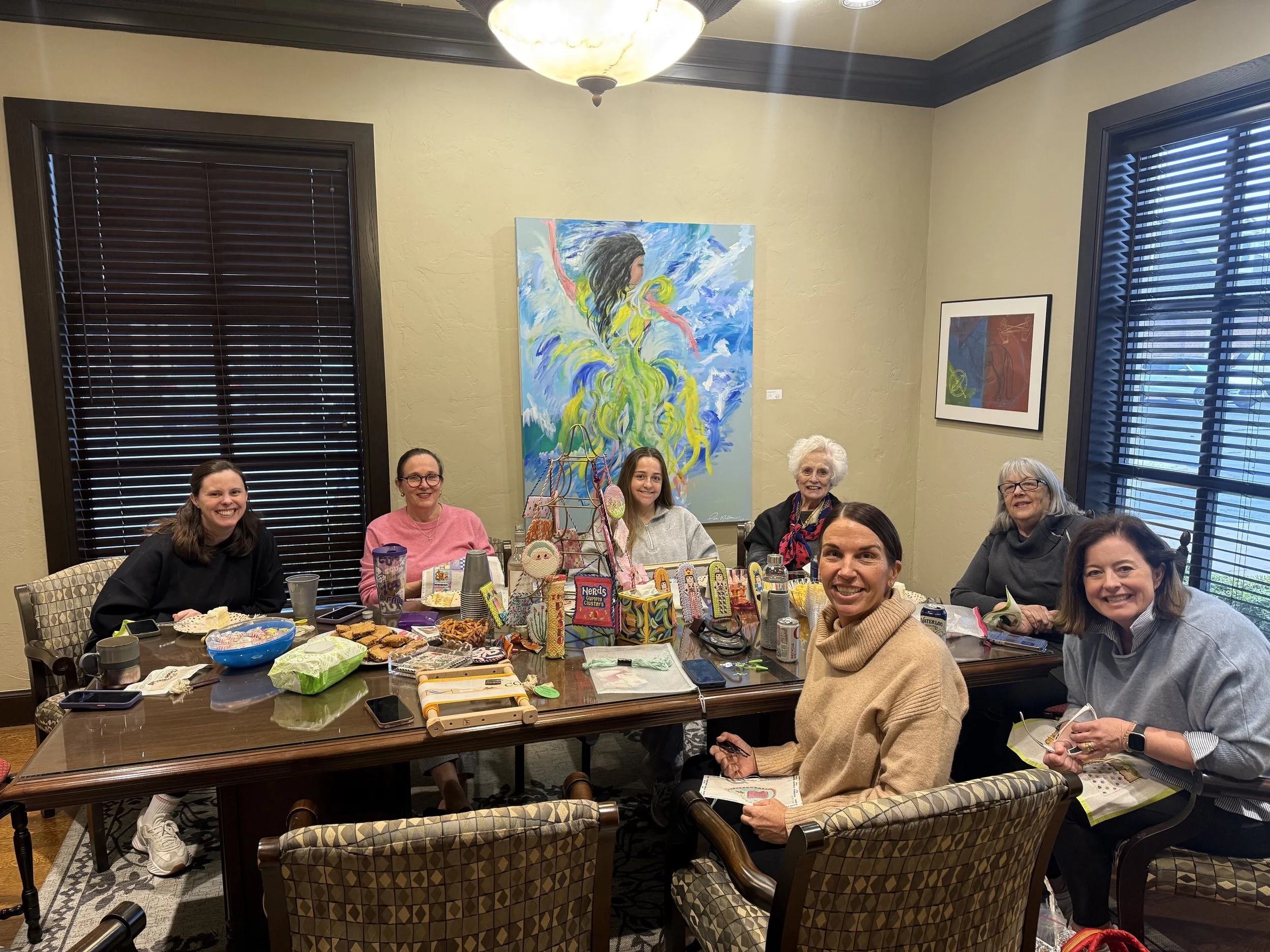 Six women smiling and sitting around a table with snacks, drinks, and holiday decorations in a room with artwork on the walls.