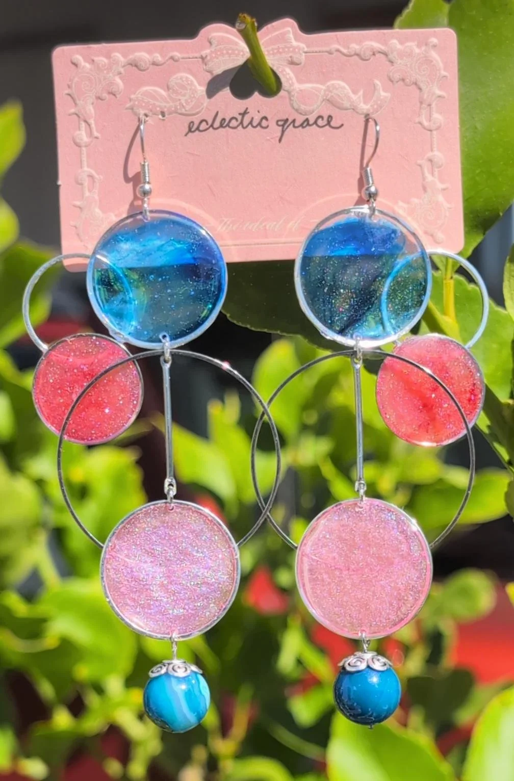 A pair of colorful dangle earrings featuring blue, pink, and glittery circular elements hanging from silver hoops, displayed on a pink decorative card with the words 'eclectic grace' written on it.
