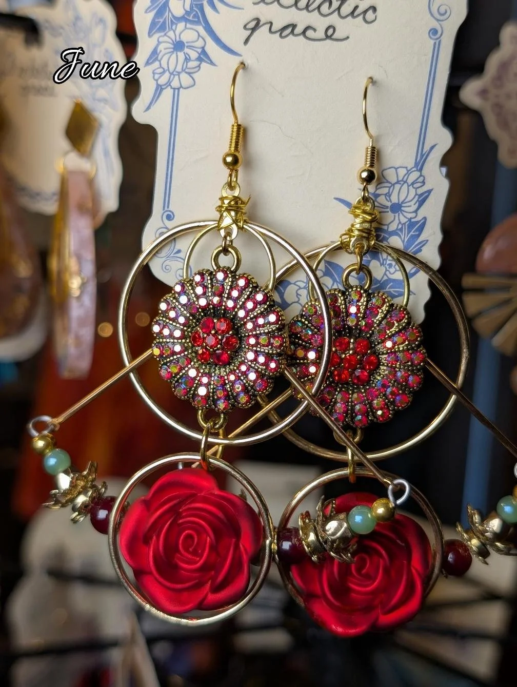 A pair of decorative earrings featuring red rose motifs, rhinestone floral designs, and multicolored beads, displayed on a card with blue floral patterns and the word 'June'.