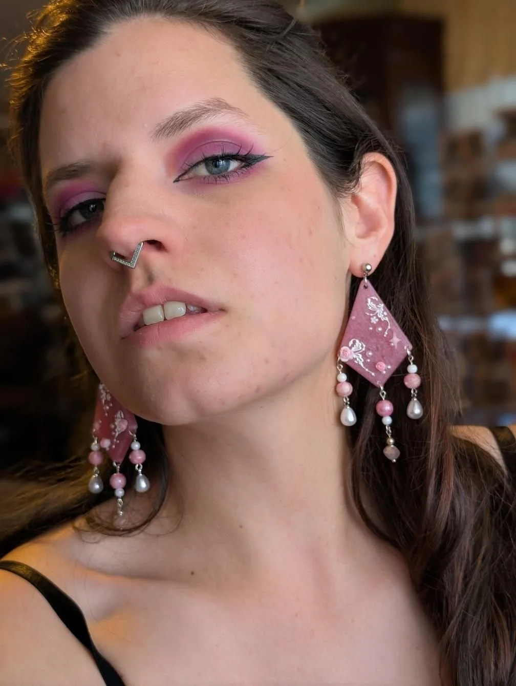 Close-up of a woman with pink eye makeup, blue eyes, a nose ring, and large pink earrings with pearls, against a blurred background.