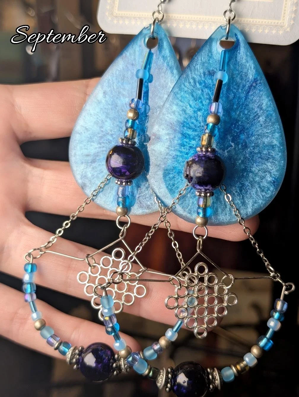 A pair of blue earrings with purple beads, chain accents, and blue rhinestone beads, displayed in front of blue teardrop-shaped leaves with "September" text.