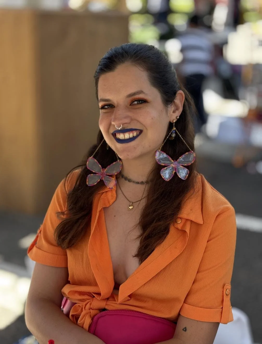 A young woman with dark brown hair, wearing a bright orange shirt and pink fanny pack, smiling with blue lipstick, sporting beaded handmade butterfly earrings, a nose septum piercing, and layered necklaces, outdoors on a sunny day.