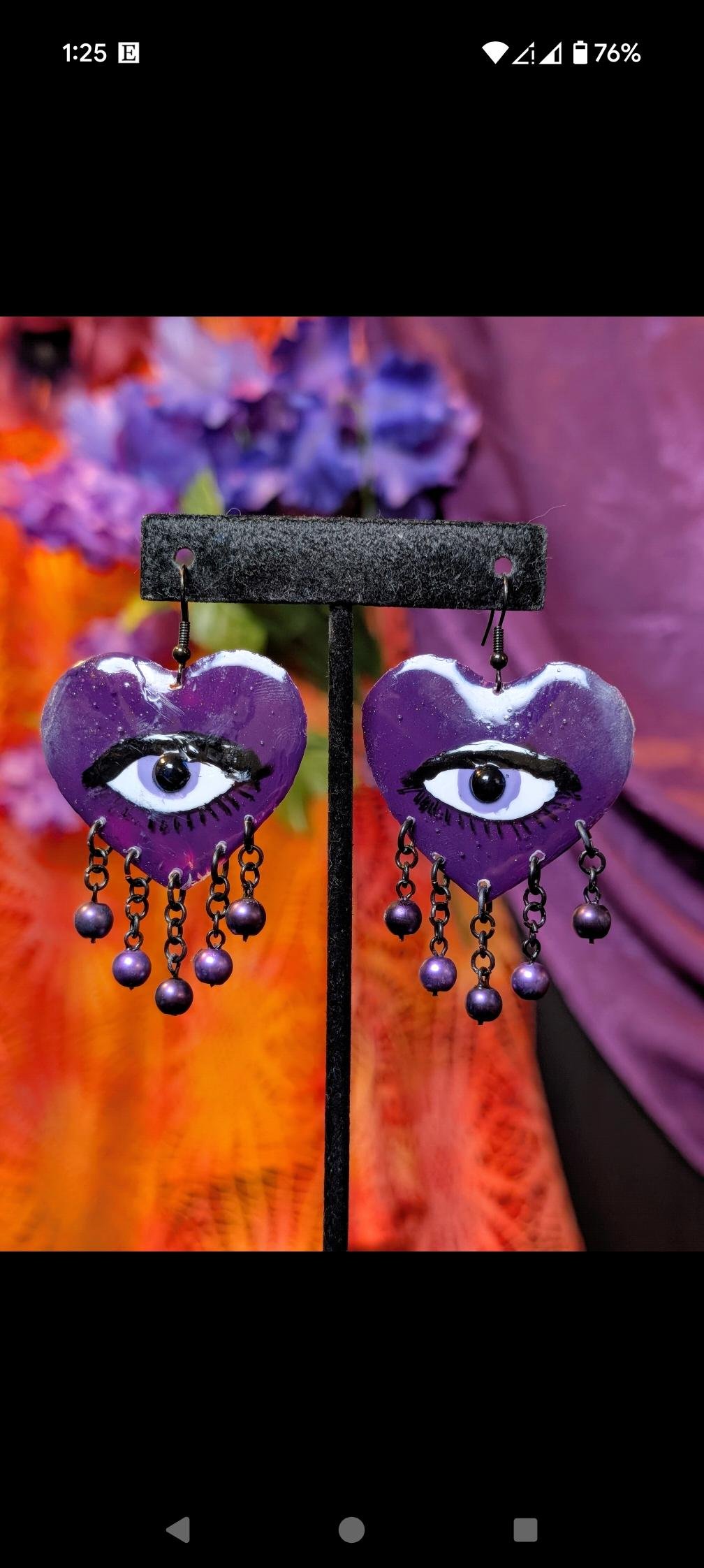 Purple heart-shaped earrings with painted eyes and dangling beads on a display stand against a vibrant background of orange, purple, and blue flowers.