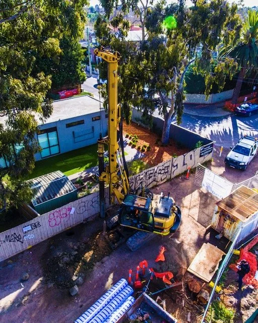An aerial view of a construction site with a yellow pile driver, surrounded by fencing, gravel, and construction materials, with trees and parked cars nearby.