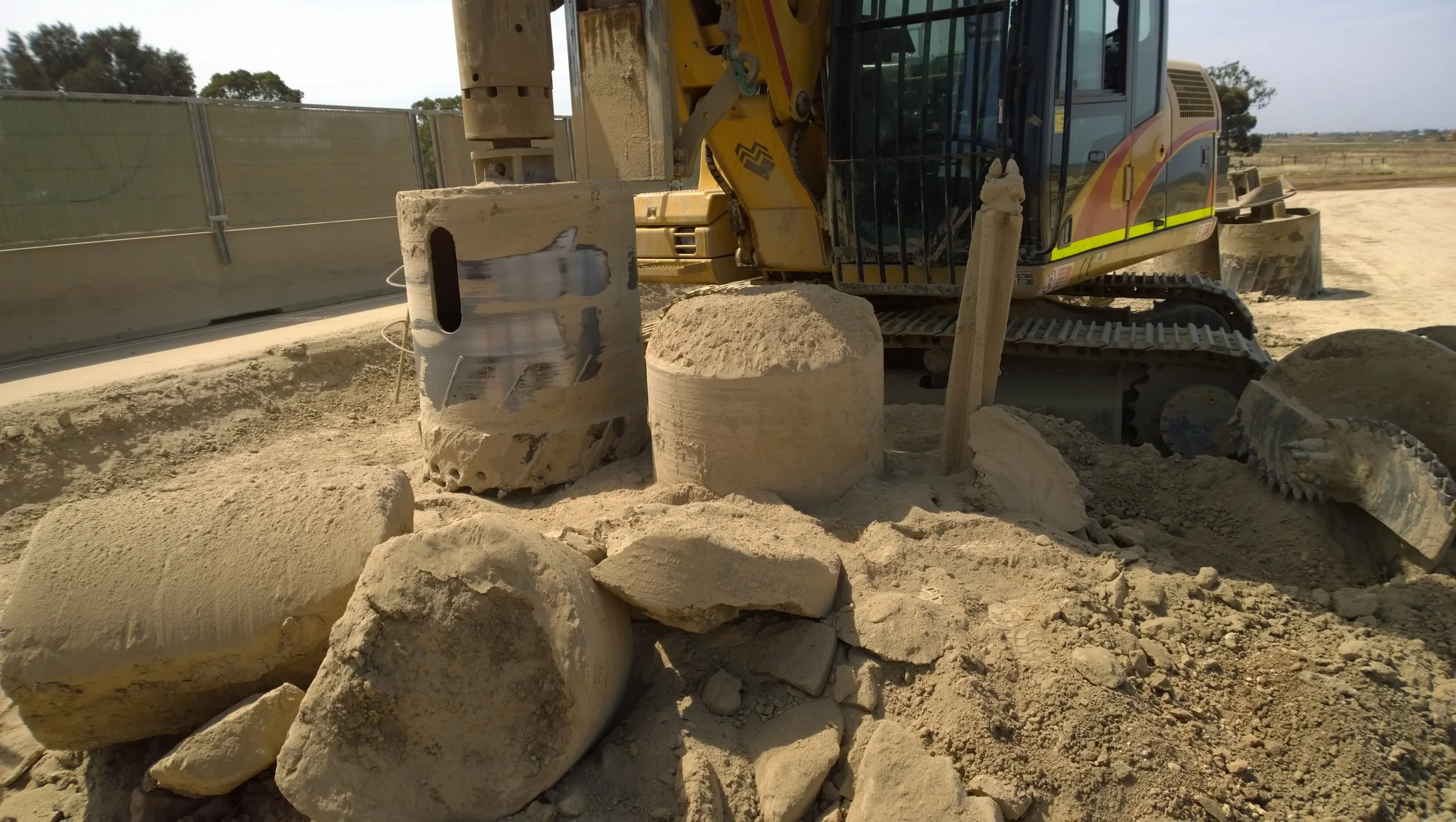 Construction site with a yellow excavator and large rocks on sandy ground.