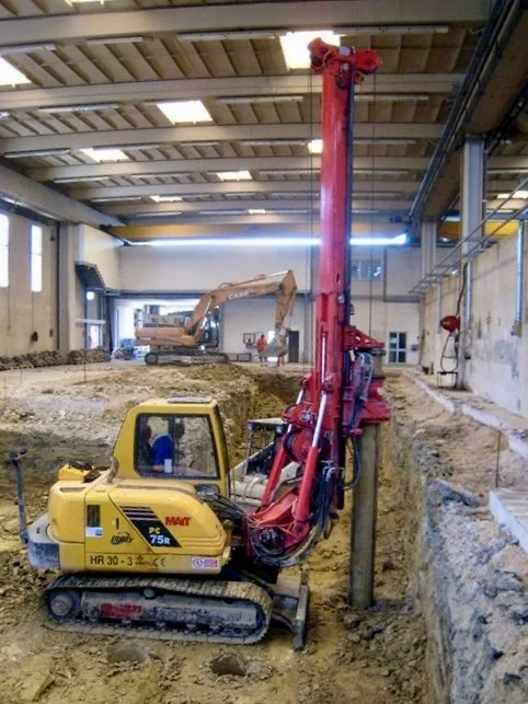 Yellow construction vehicle with a tall red drilling rig inside a large unfinished building, surrounded by dirt and construction equipment.