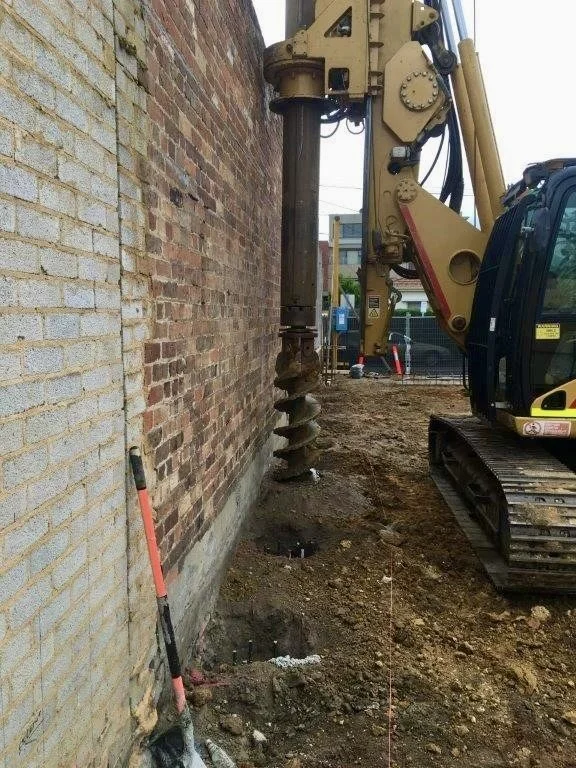 Construction site with an excavator drill attached to a yellow arm, drilling into a brick wall. The ground has holes and dirt, and a shovel or pickaxe is leaning against the wall.